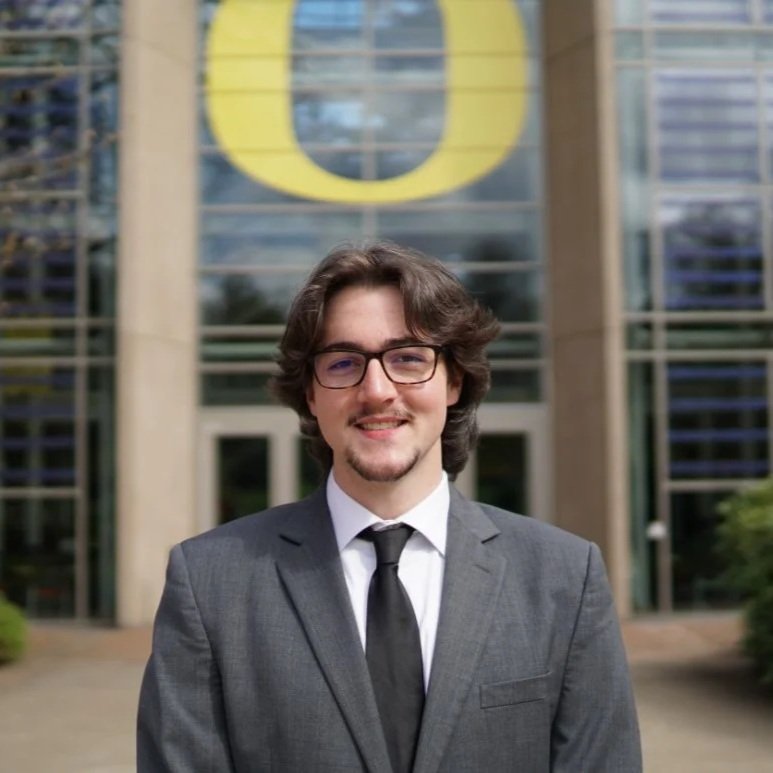 Young man in a gray suit and black tie standing outside modern building with glass windows and a large yellow 'O' logo in the background.