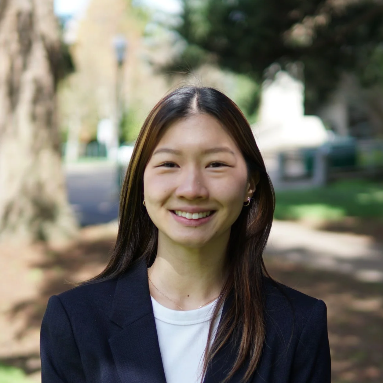 A young woman smiling outdoors in a park setting with trees and a pathway.