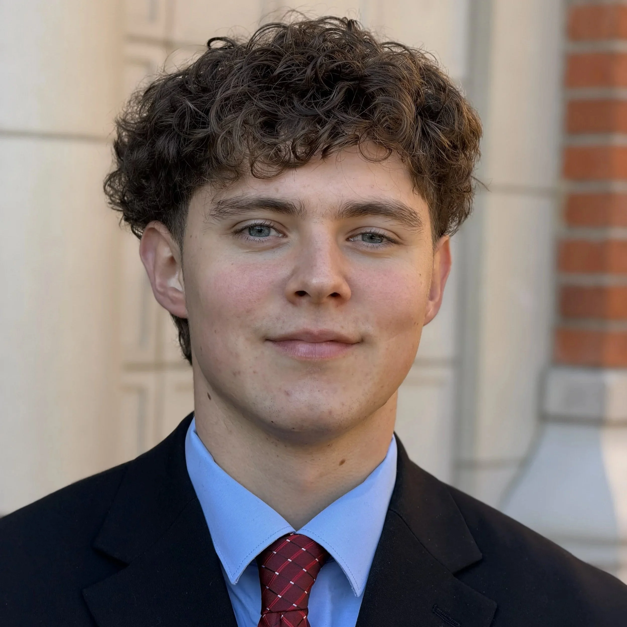 A young man with curly brown hair, wearing a black suit, blue shirt, and red tie, standing outside with a brick building and lamp in the background.