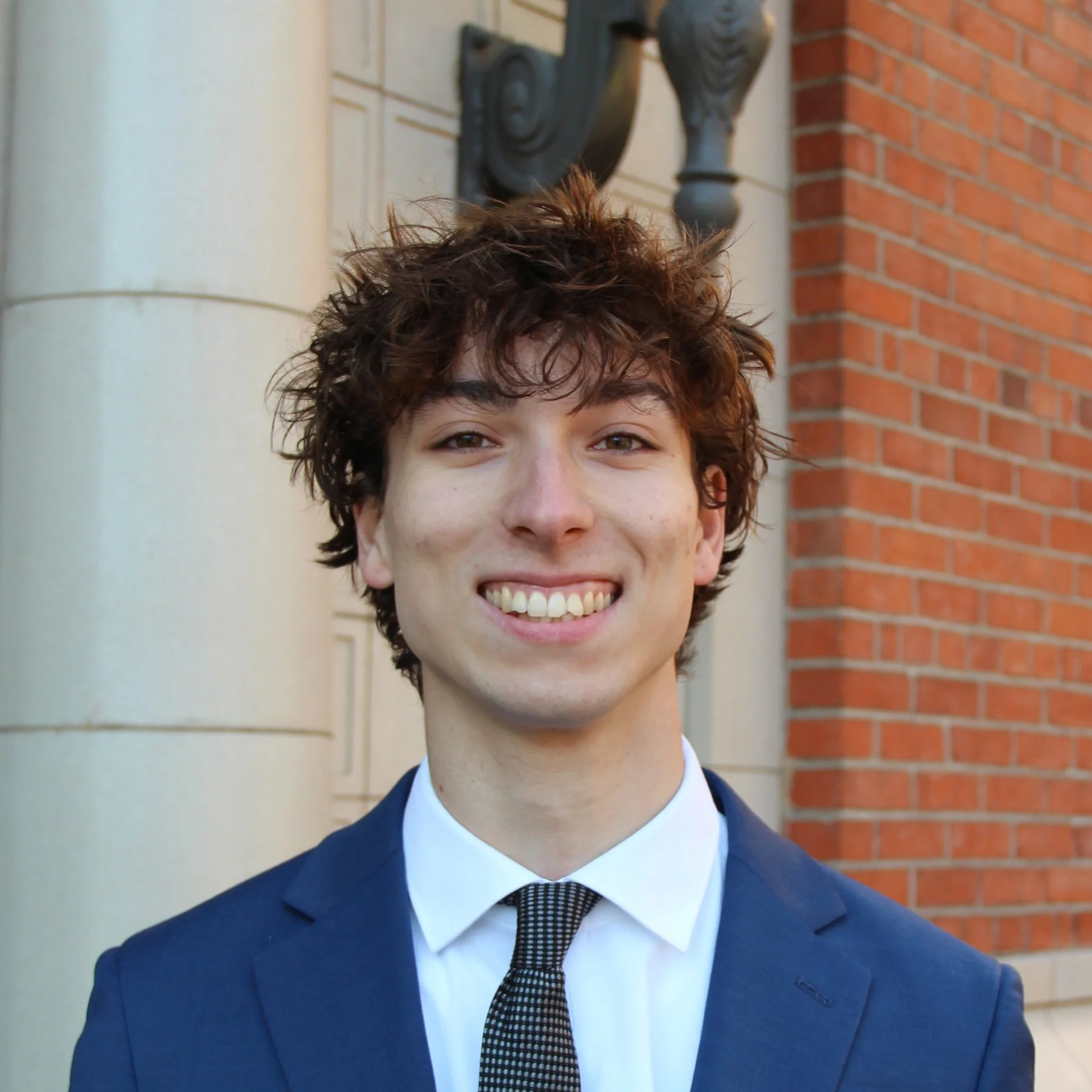 Young man with curly brown hair smiling, wearing a navy blue suit, white shirt, and black patterned tie, standing outdoors in front of a brick building and columns.