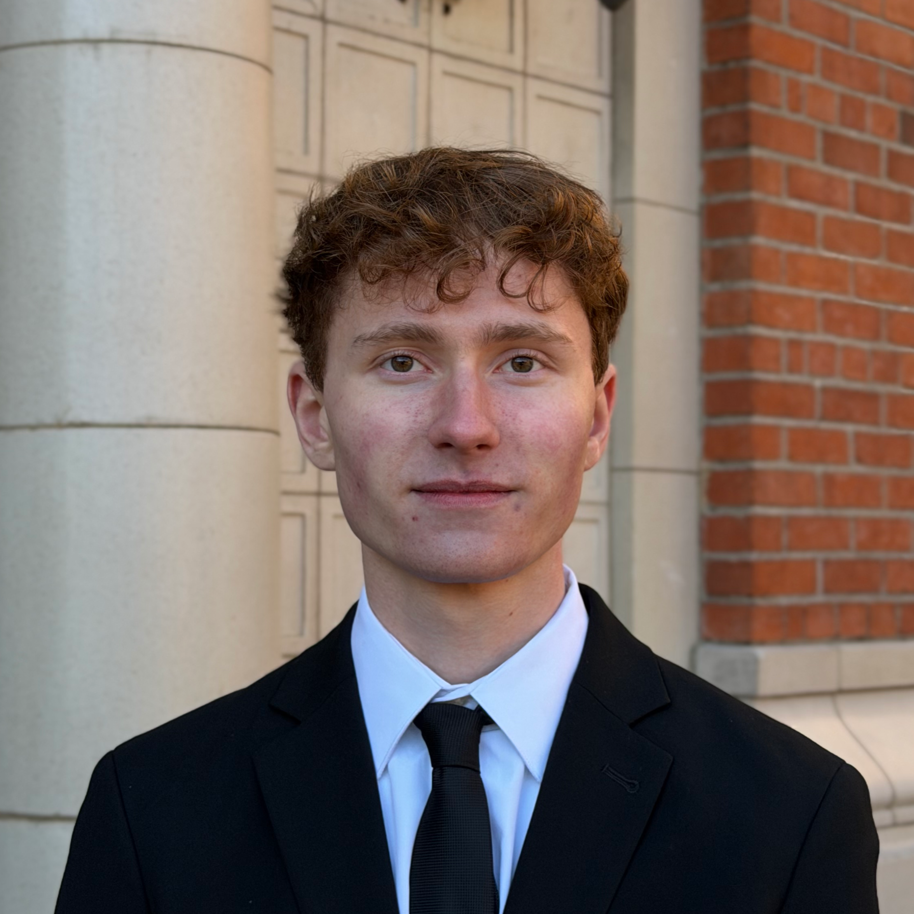 A young man with curly brown hair and fair skin wearing a black suit, white shirt, and black tie standing in front of a building with brick and stone walls.