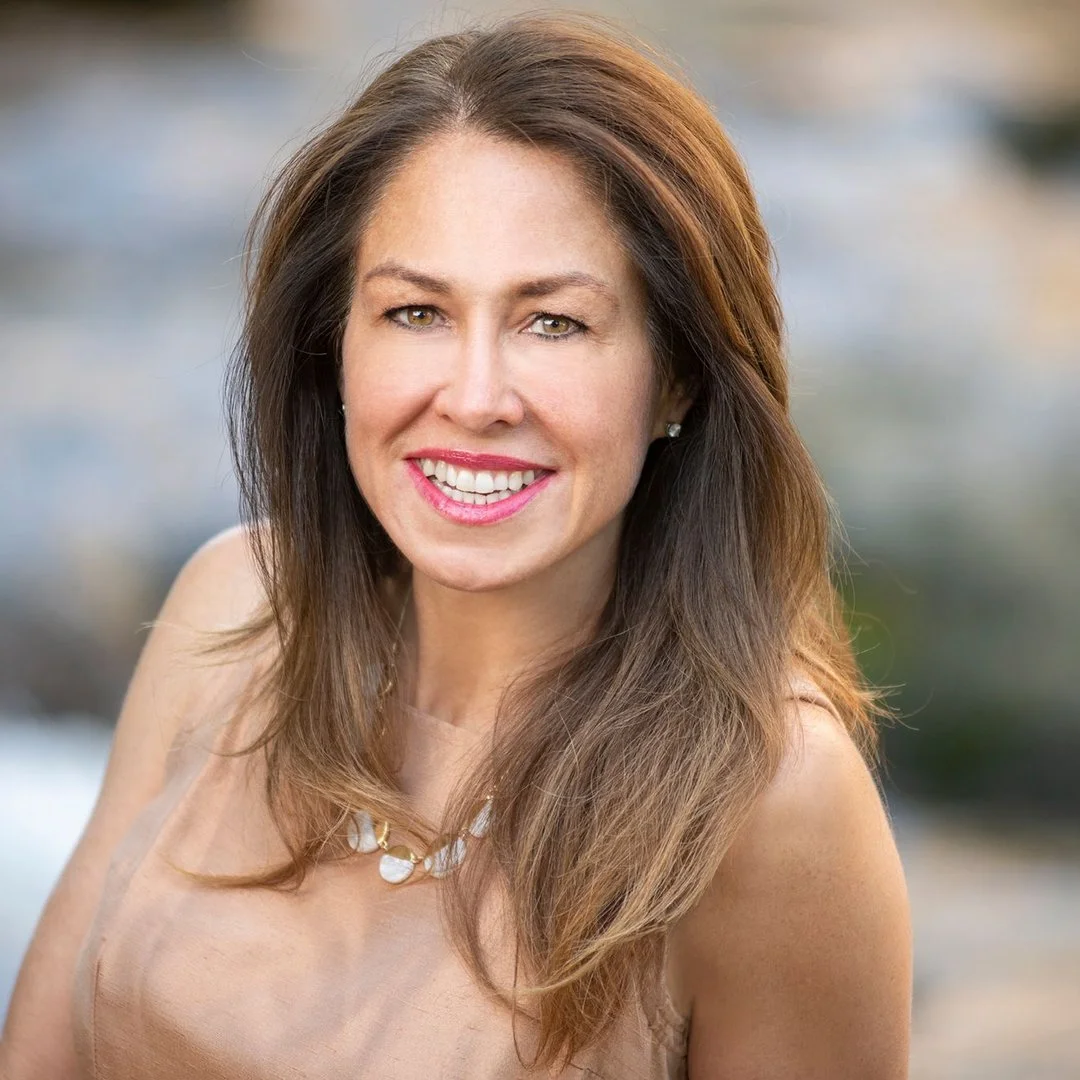 A woman with long brown hair smiling outdoors with a blurred natural background.