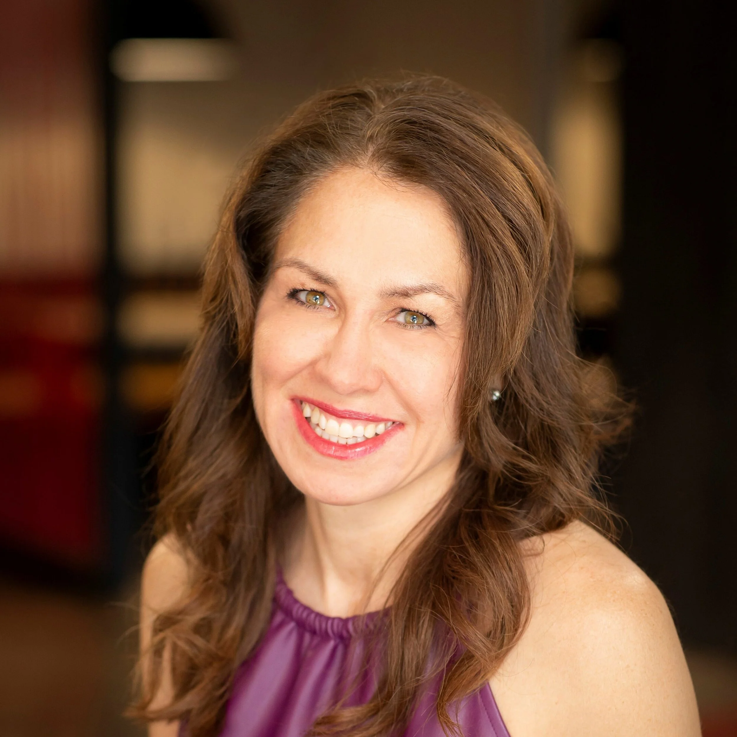 A woman with shoulder-length brown hair, smiling with white teeth, wearing a purple sleeveless top, indoors with a blurred background.