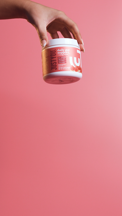 Hand holding a container of Just Sty.io daily gut support supplement against a pink background.