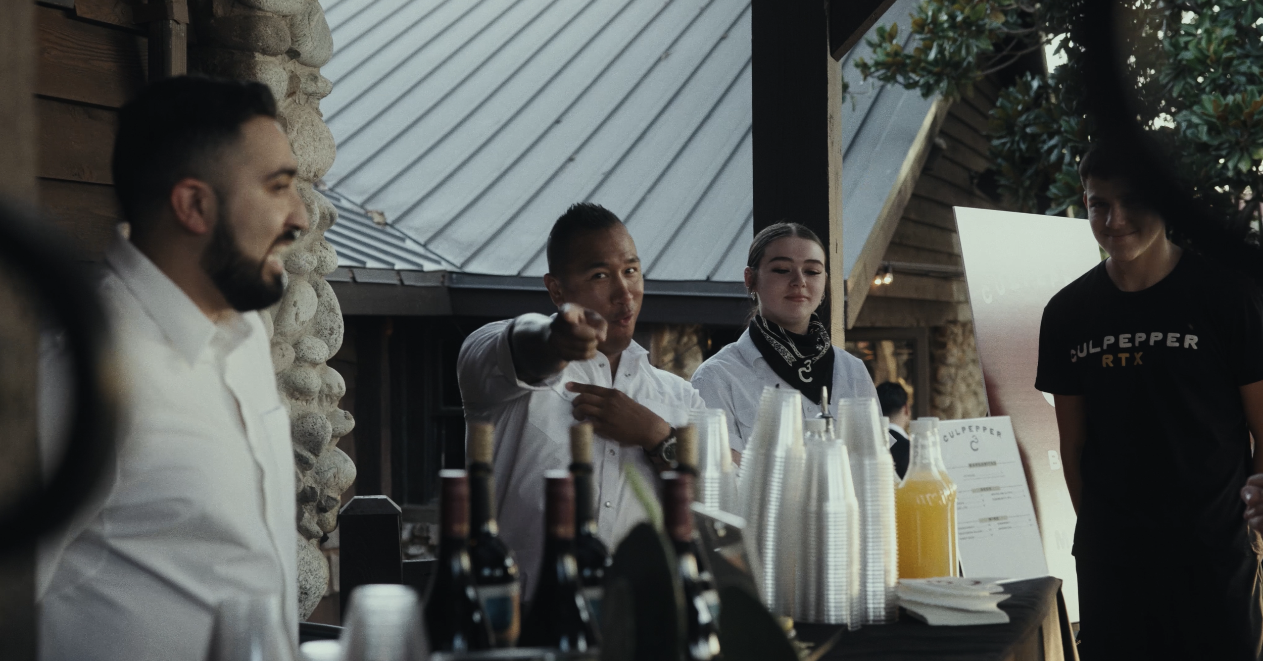 Four people behind a bar or counter, with bottles and cups, at a restaurant or event.