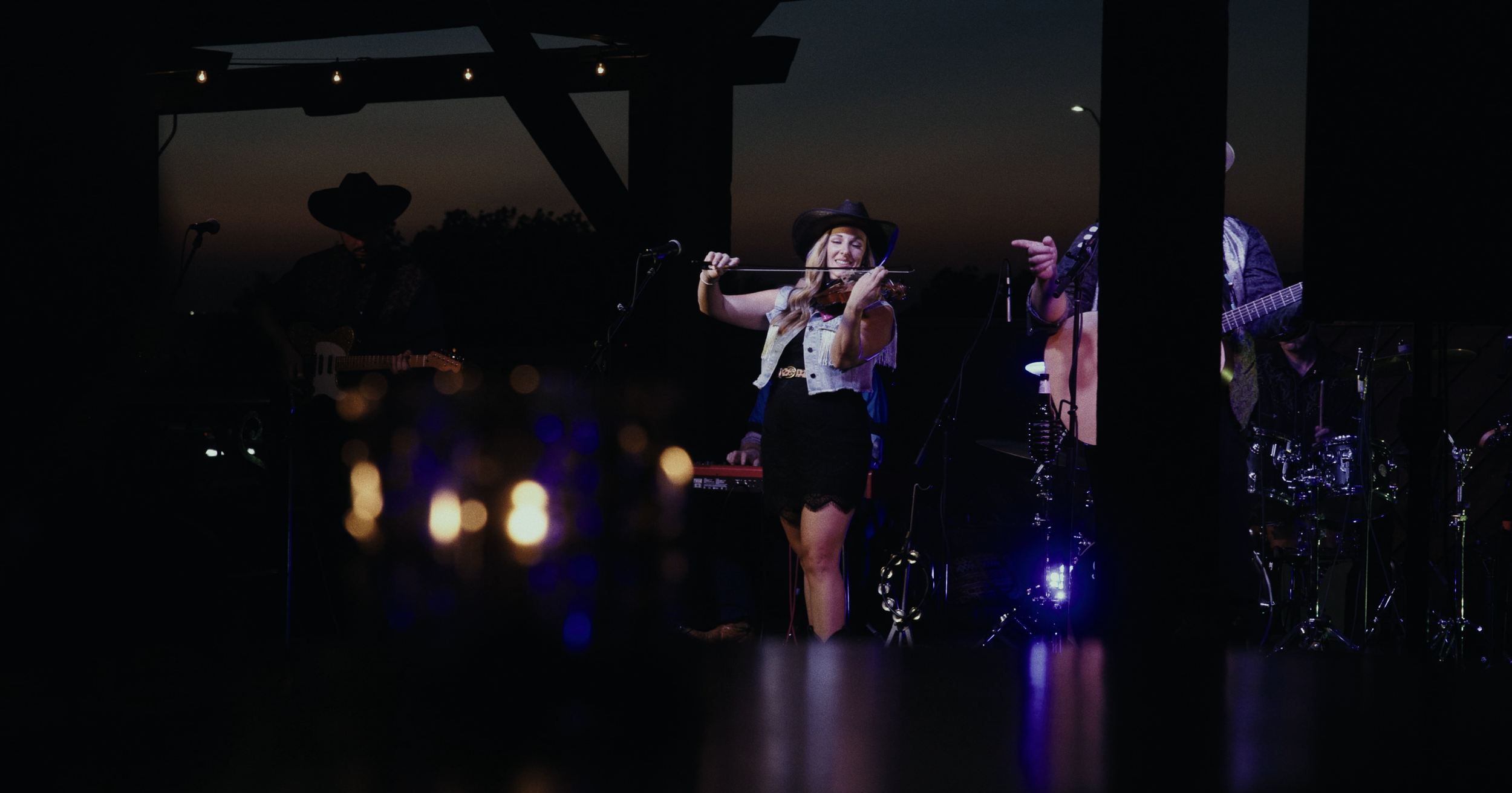 Musician playing violin on stage during a nighttime outdoor concert with band members and sunset in background.