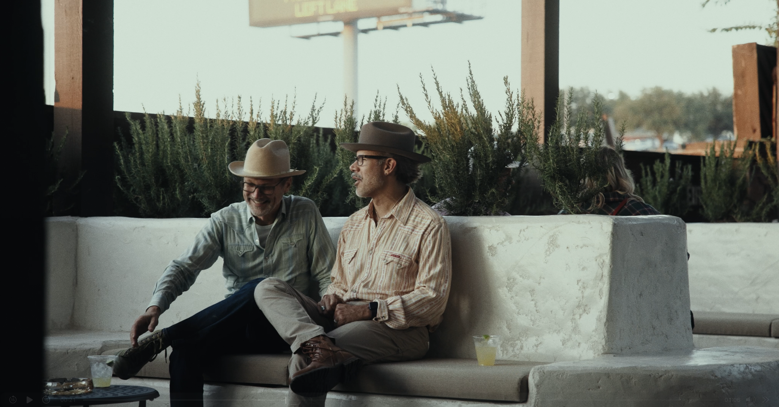 Two men sitting on a white marble bench, smiling and talking. One man is wearing a beige hat, glasses, and a striped shirt; the other man is wearing a brown hat, glasses, and a plaid shirt. A drink with a lime wedge is on the bench next to them, and 