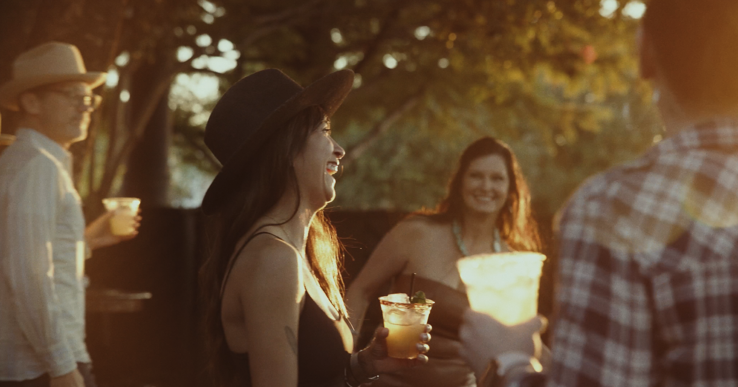 People enjoying drinks at an outdoor gathering during sunset, wearing casual summer outfits and hats, with trees in the background.