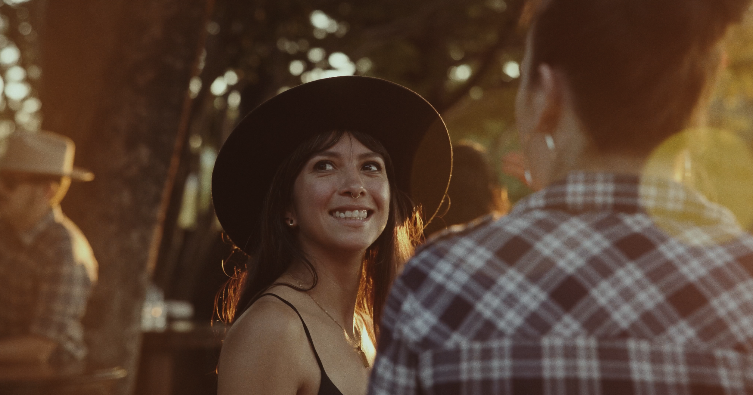 A woman with long dark hair, wearing a wide-brimmed black hat and a black spaghetti strap top, smiling and looking at a person in a checkered shirt, outdoors during sunset.