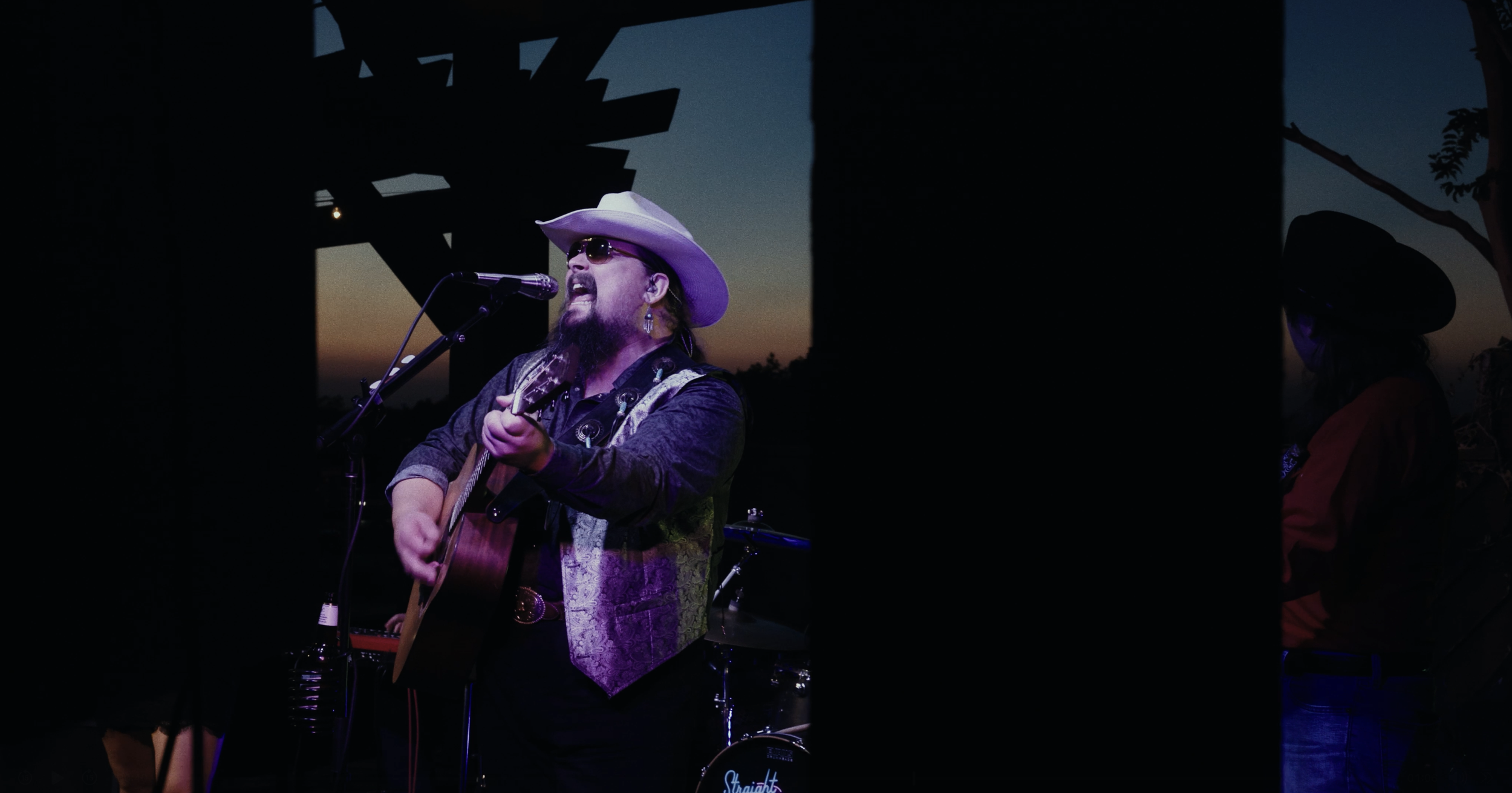 Male musician wearing a white cowboy hat, sunglasses, and a patterned vest singing into a microphone while playing an acoustic guitar during an outdoor performance at sunset.
