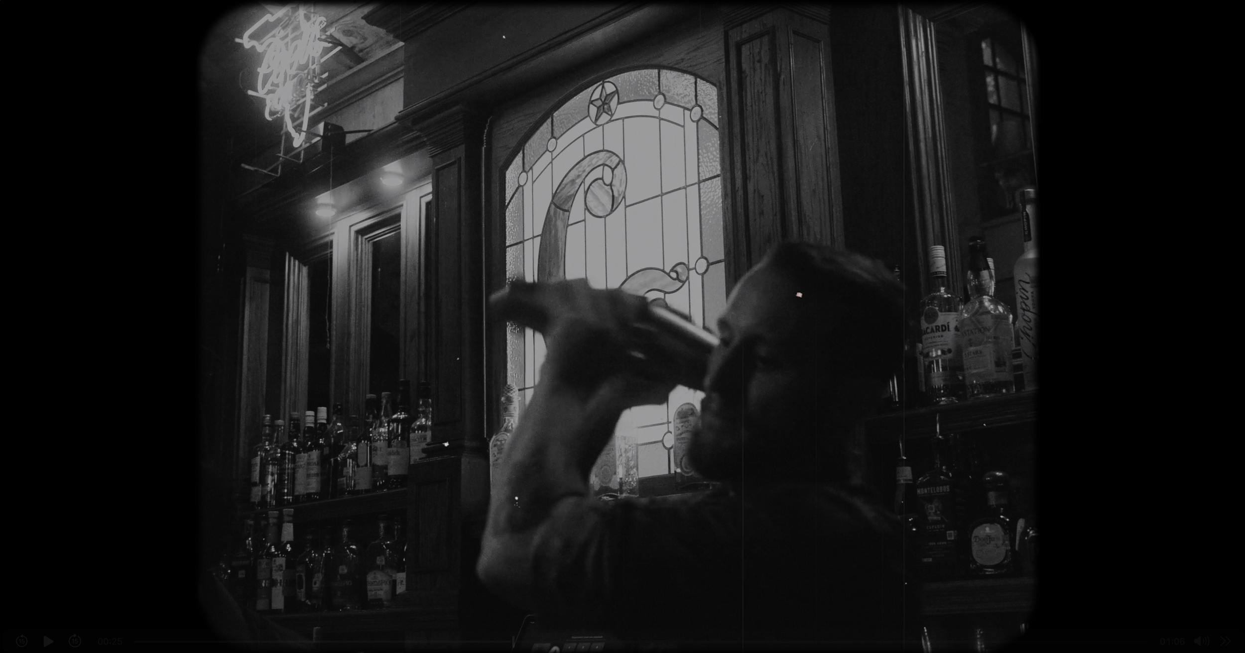A person drinking from a bottle at a bar with bottles of alcohol on shelves behind them, stained glass window, and neon sign partially visible.