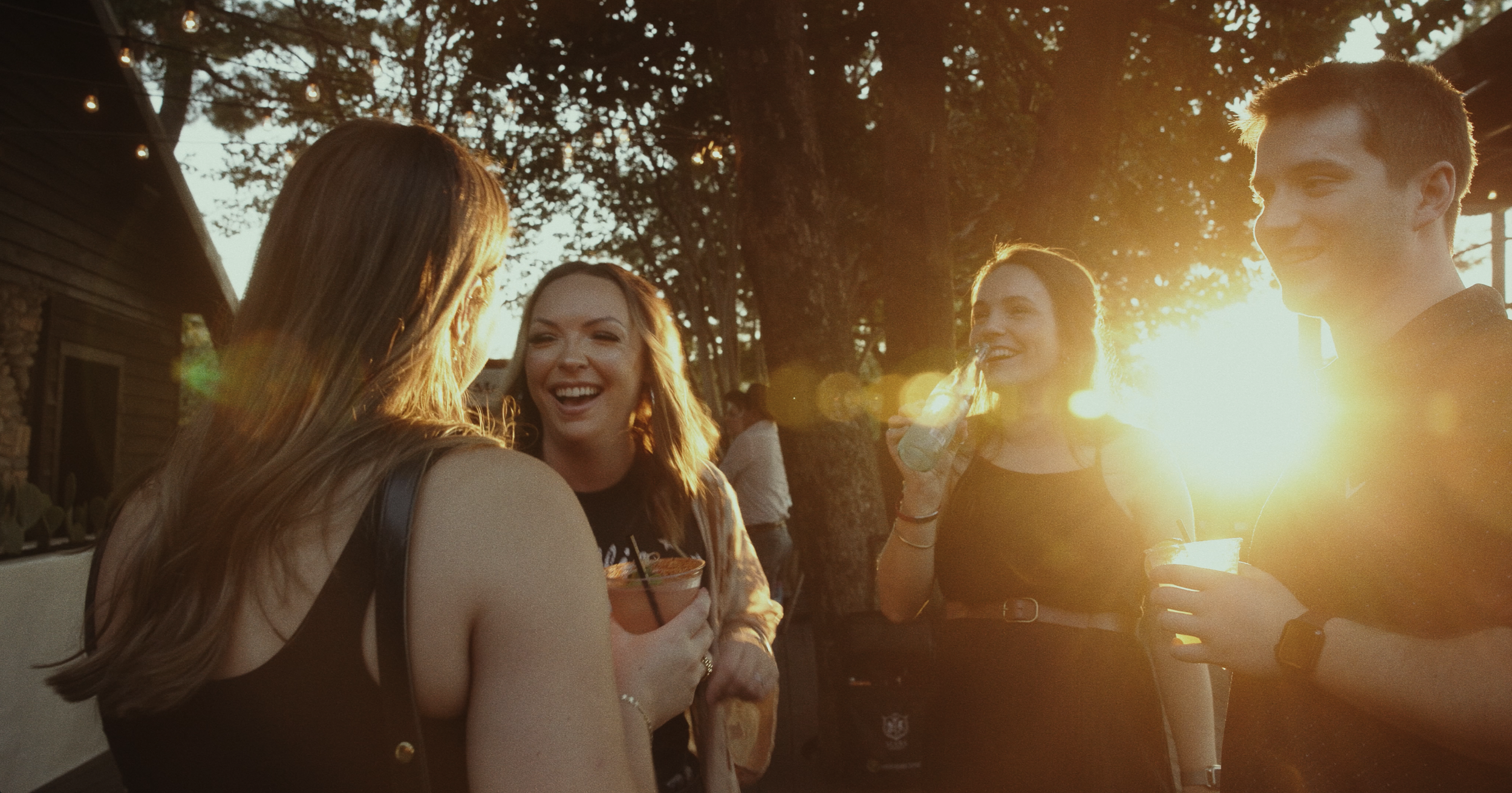 Four young adults, three women and one man, enjoying drinks and laughing outdoors during sunset. They are gathered closely, with trees and string lights in the background.