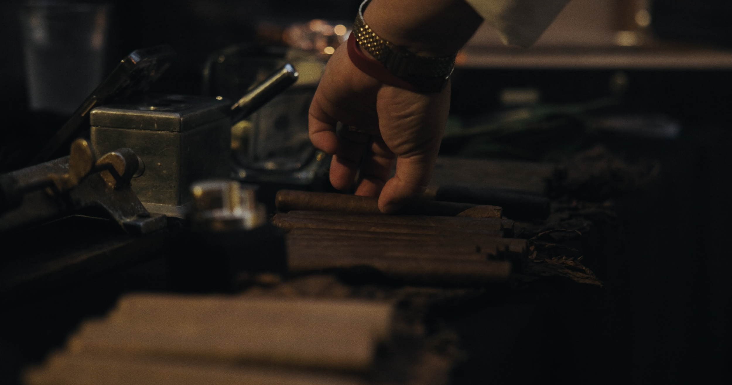 A person's hand arranging wooden sticks or tools on a table, with various small objects and metal tools around in a dimly lit setting.