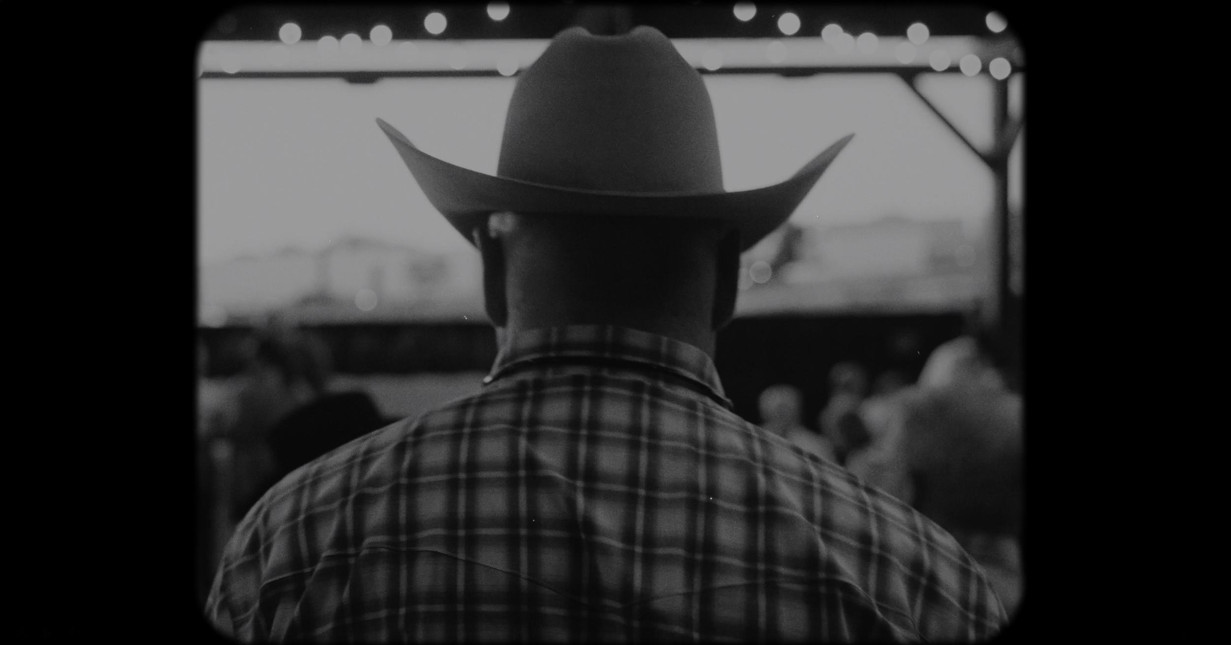 A black and white photograph of a man wearing a cowboy hat and a plaid shirt, viewed from behind, at an outdoor event or gathering with blurred people and structures in the background.