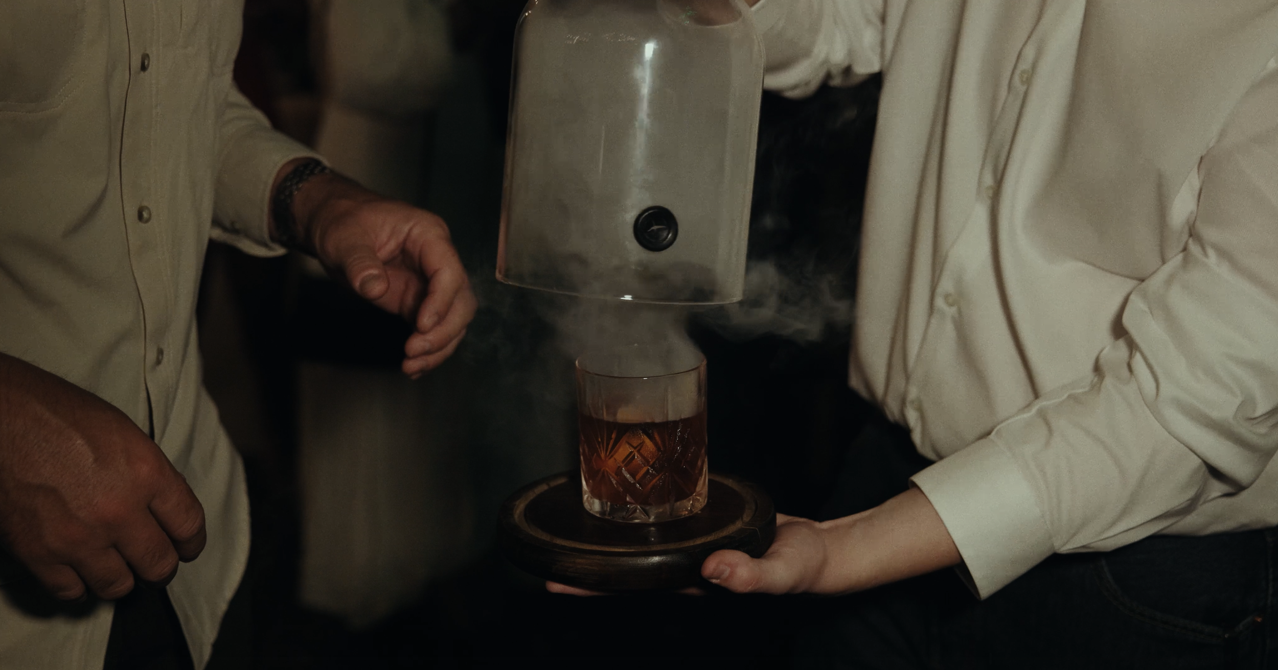 Two people serving a drink from a glass teapot into a glass on a wooden tray in a dimly lit setting.