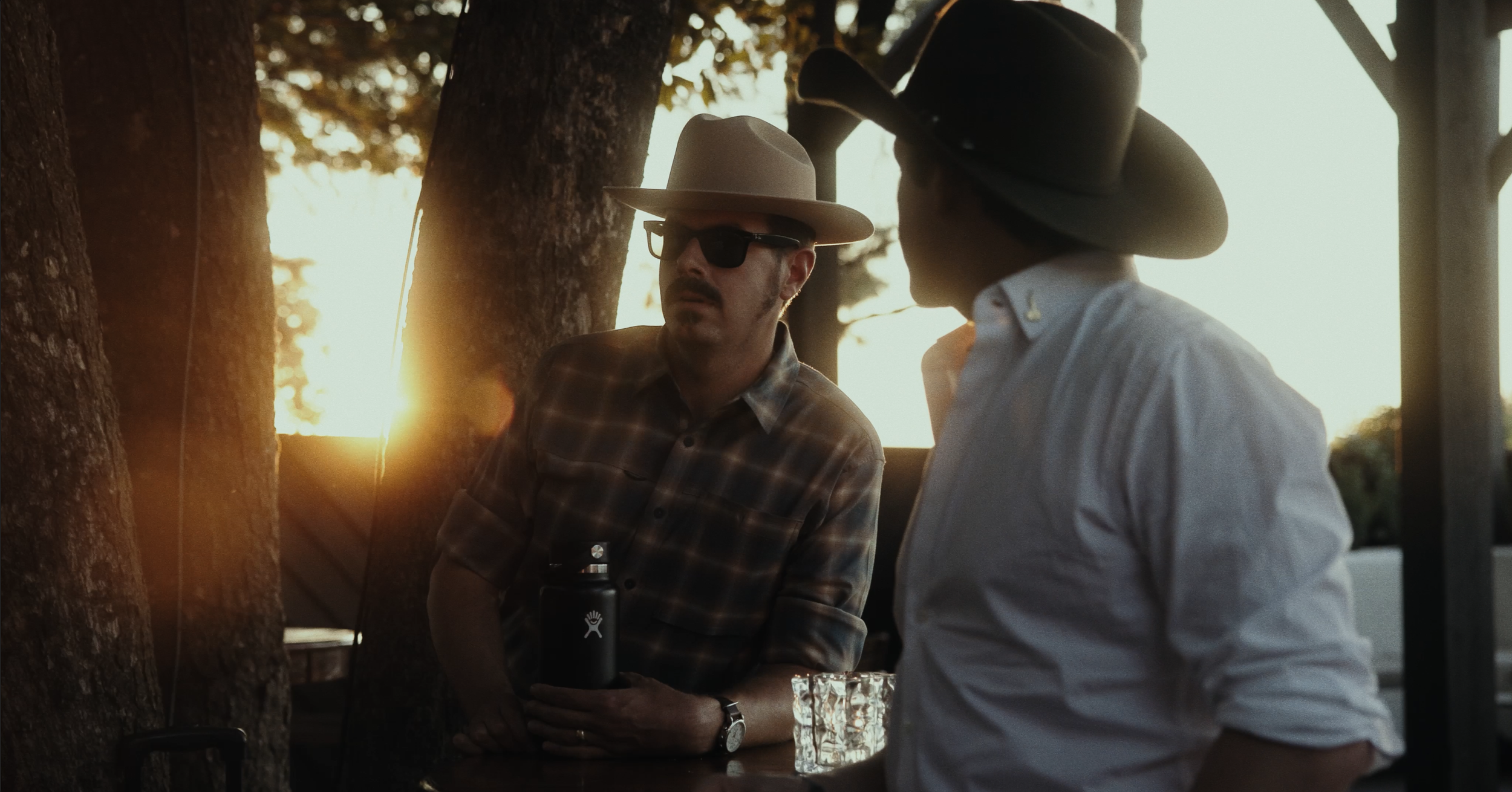 Two men in western attire wearing wide-brimmed hats, sitting outdoors during sunset. One man with sunglasses is holding a black water bottle, and a clear glass is on the table in front of him.