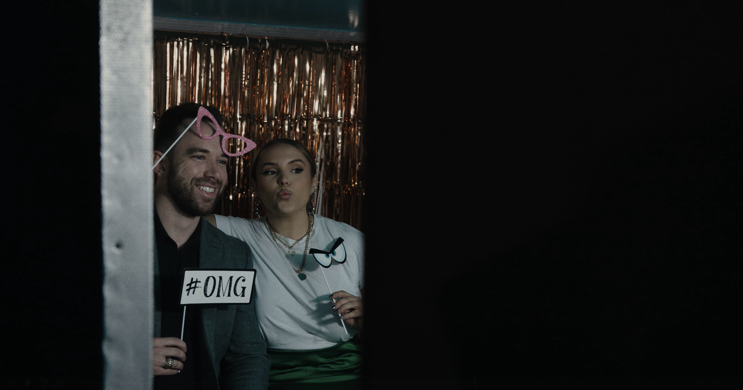 Two people at a photo booth, one man and one woman, holding signs, with fun glasses prop, in front of a shiny, metallic curtain background.