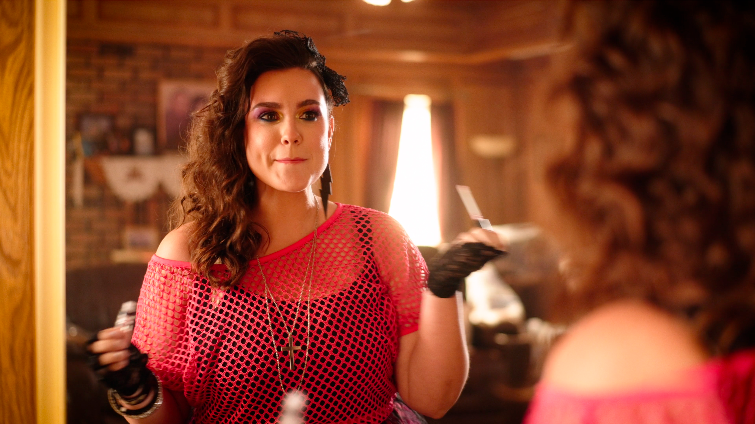 A young woman with curly brown hair looks at herself in a mirror, adjusting her fishnet pink top. She is in a warmly lit room with a brick wall and curtains in the background.