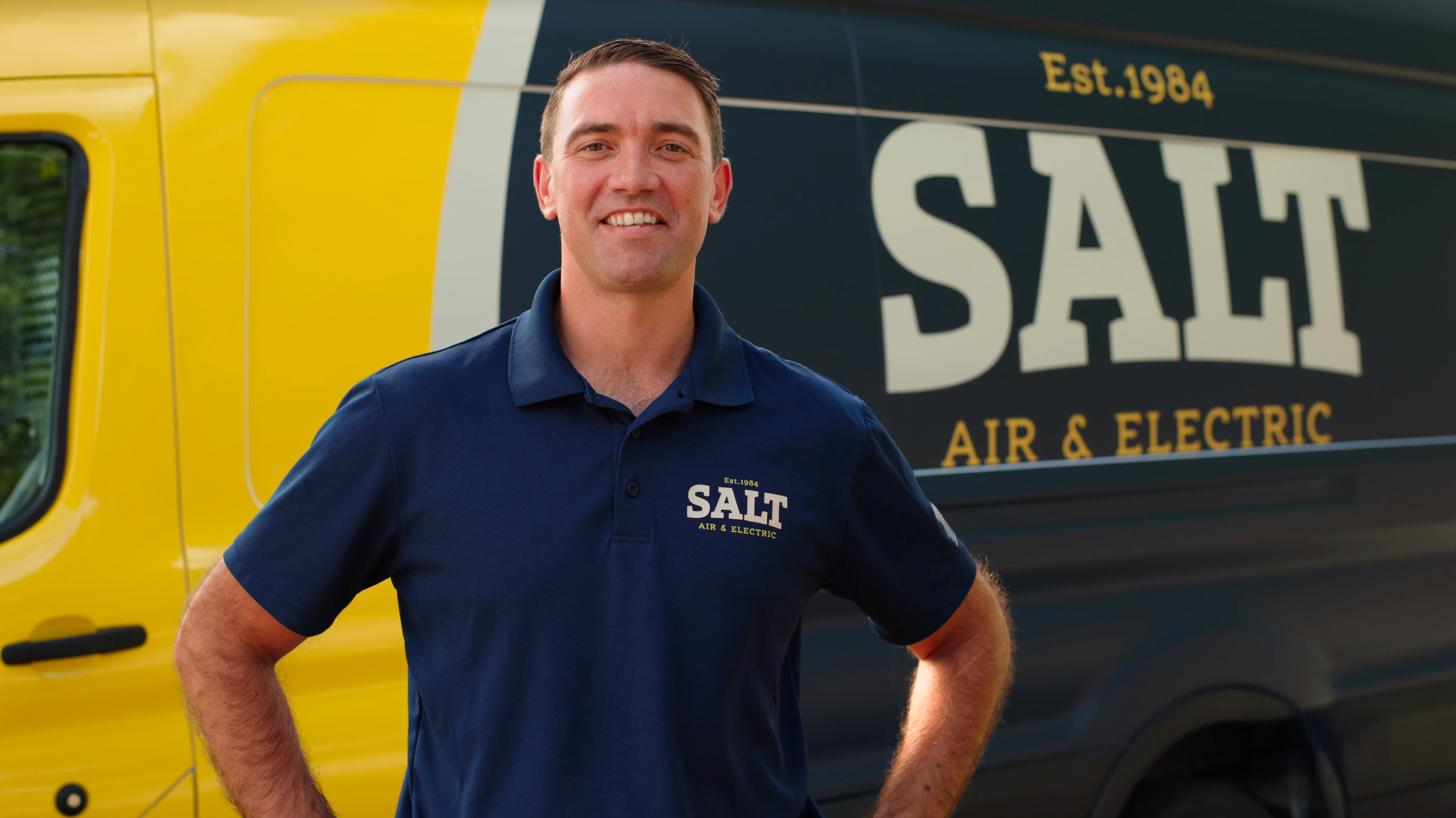 A smiling man in a navy blue polo shirt standing in front of a yellow and black Salt Air & Electric service van.