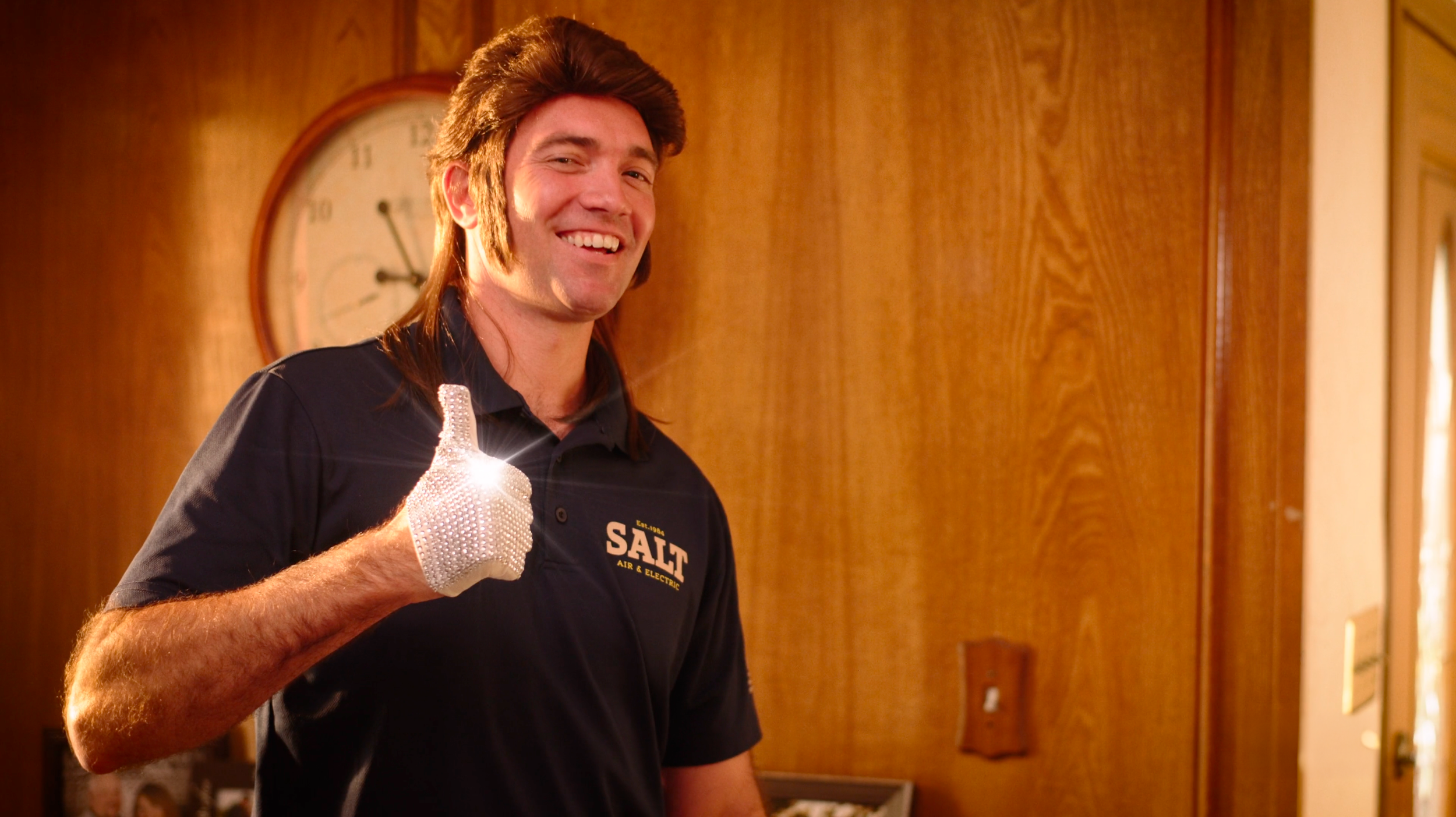 A smiling man wearing a black polo shirt with 'Salt Air & Electric' logo, giving a thumbs-up with a sparkly glove in a room with wooden walls and a clock in the background.