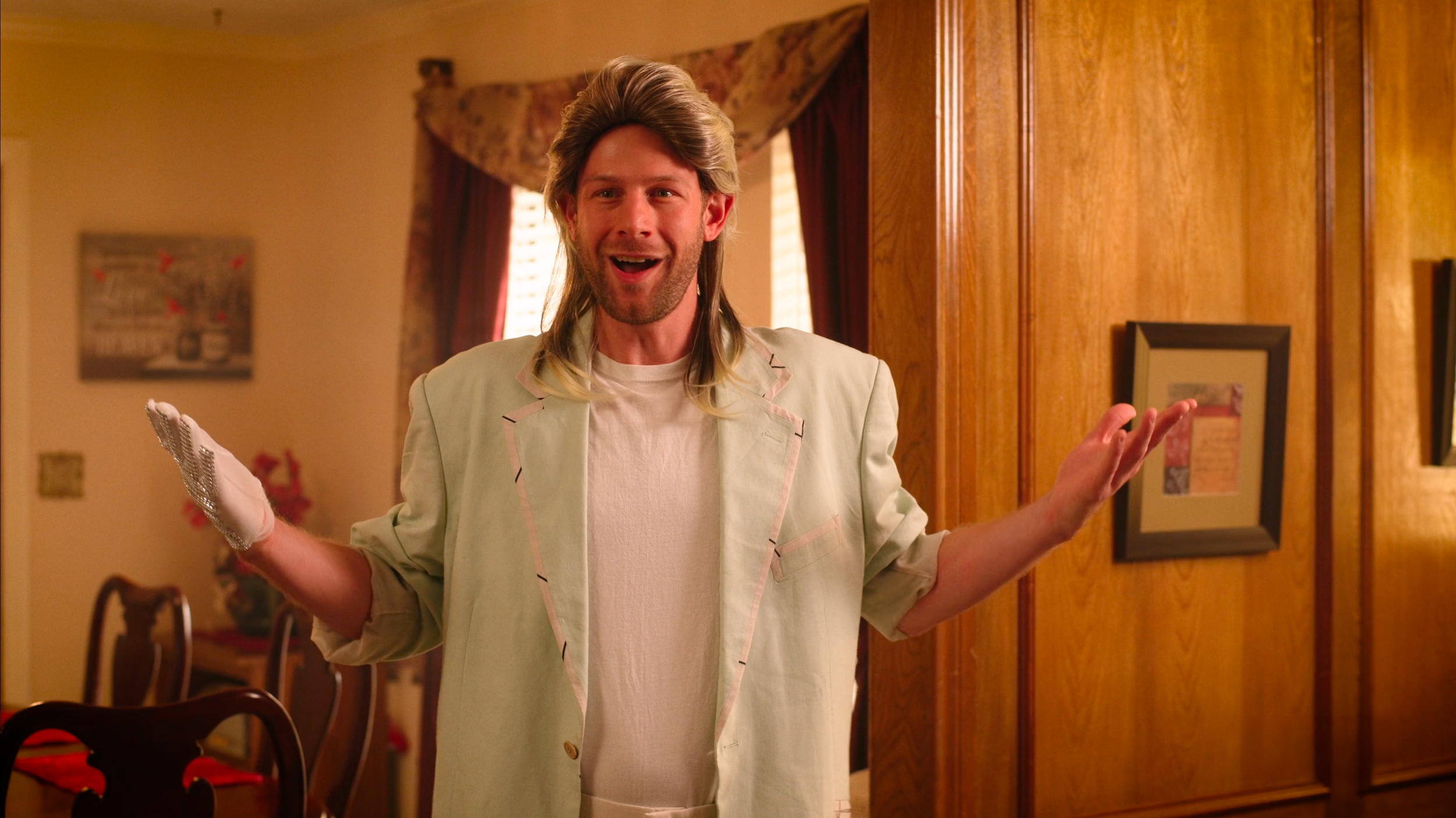 A man with long hair and a beard, wearing a light-colored blazer and shirt, smiling with his hands raised in a questioning or playful gesture, standing in a warmly decorated room with wooden walls and framed pictures.
