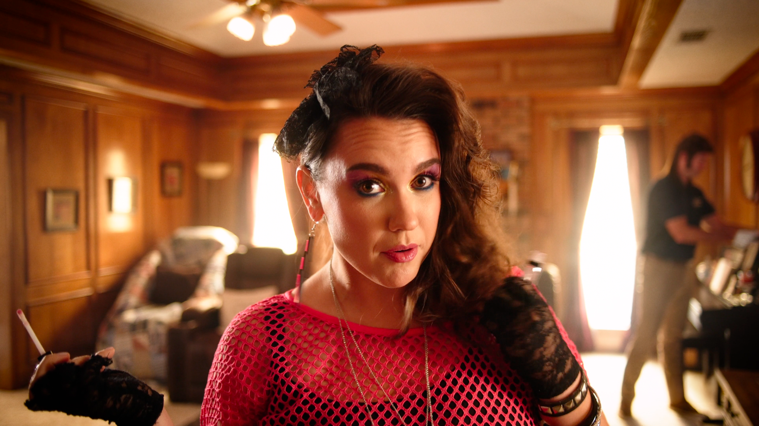 Woman with curly brown hair and colorful makeup wearing a pink fishnet top and black lace gloves, looking at camera in a wood-paneled living room with a blurred man in the background.