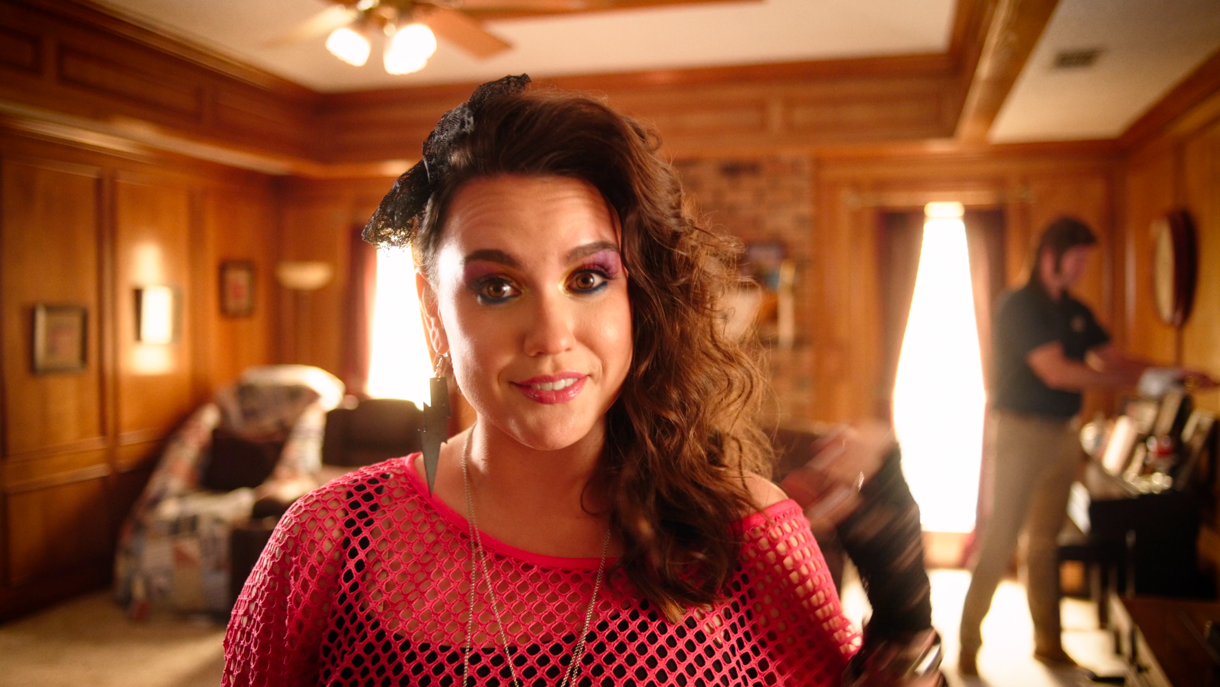 A woman with curly hair, wearing colorful makeup and a pink crochet top, standing in a cozy wood-paneled room with a man in the background.
