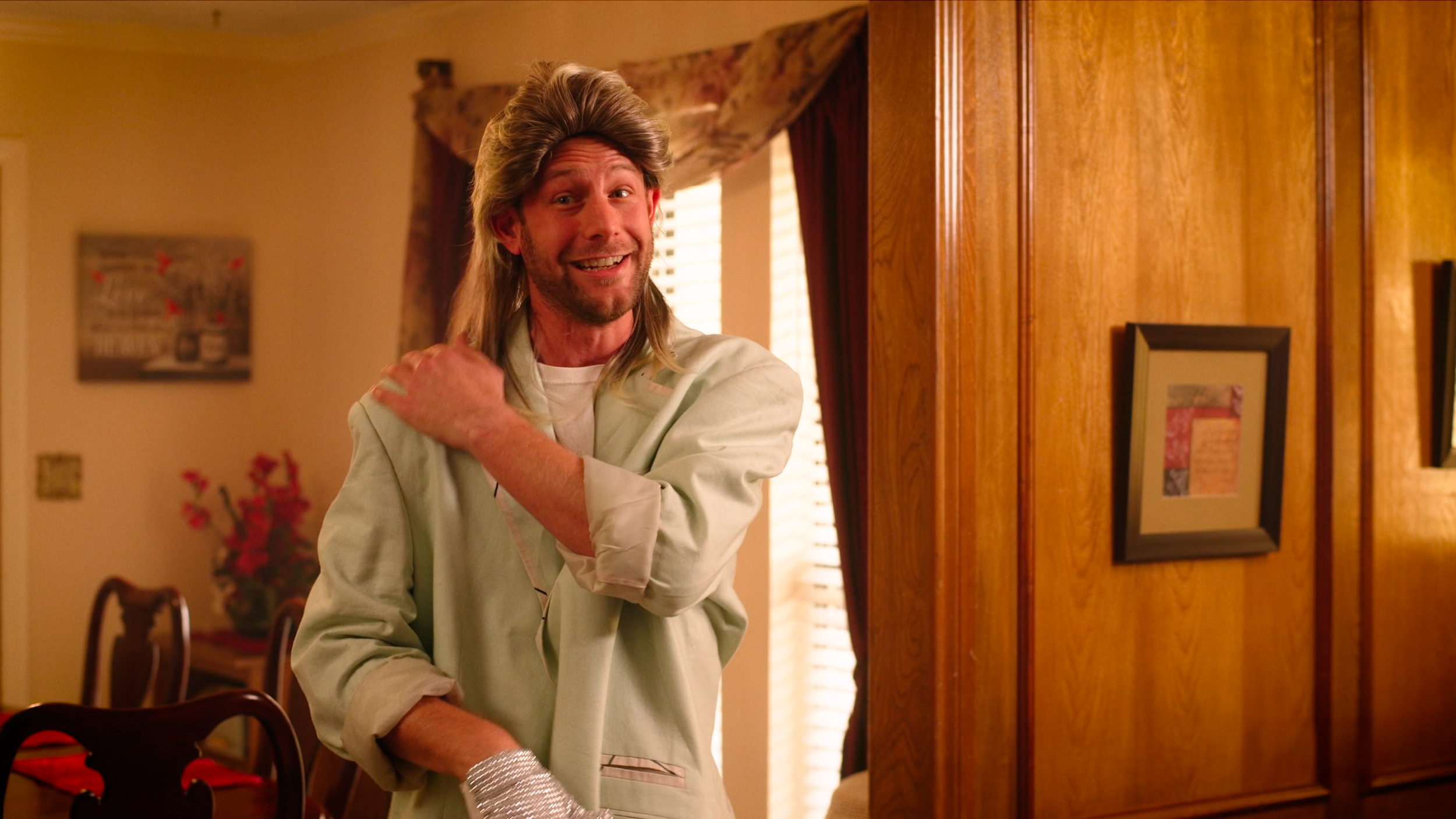 A man with a mullet hairstyle smiling and adjusting his shirt in a warmly lit dining room with wooden paneling and framed pictures on the wall.