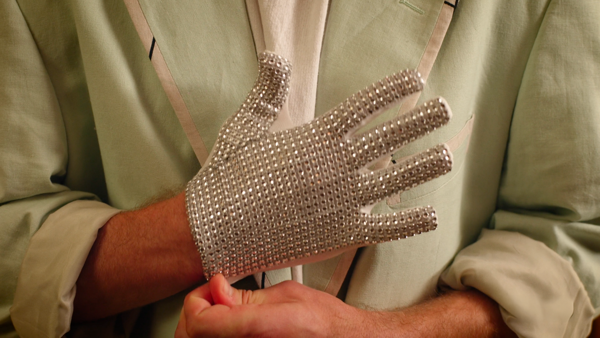 A person wearing a vintage light green suit with rolled-up sleeves holding a glittering rhinestone glove.