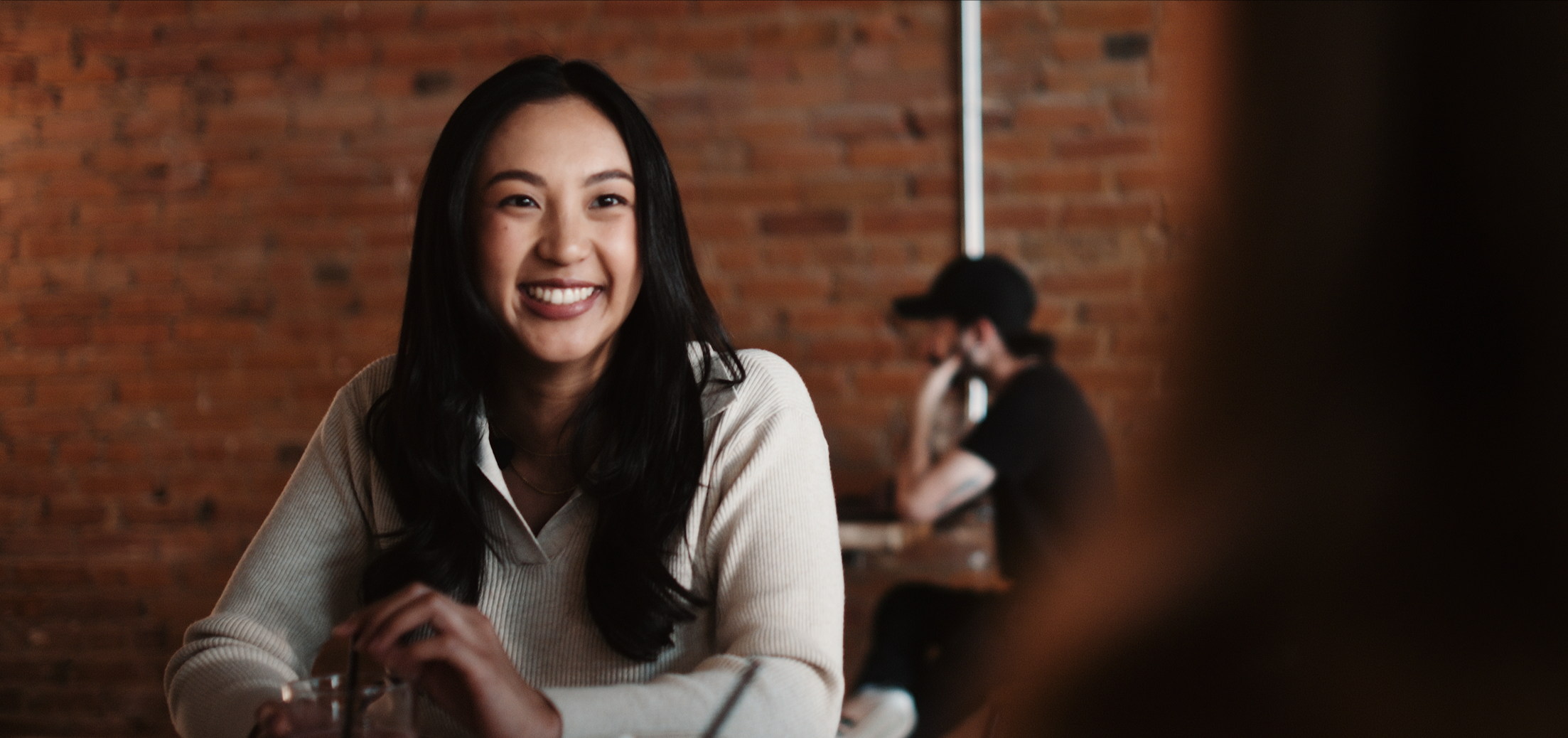 A smiling young woman with long black hair sitting at a table in a cafe with a brick wall background.