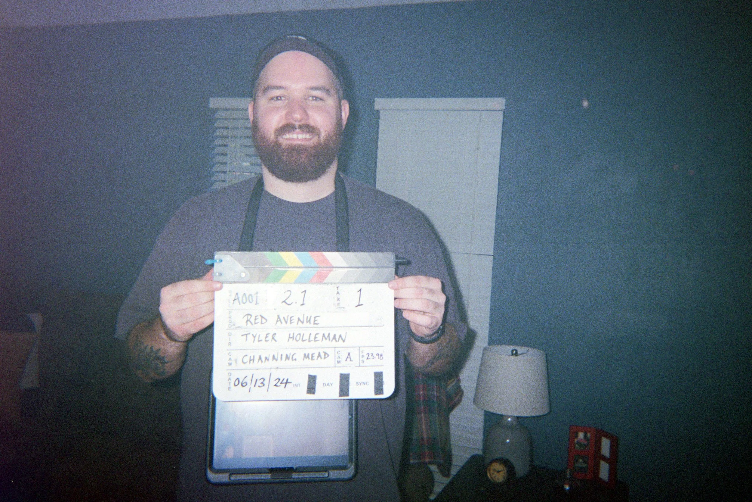 Man with a beard holding a clapperboard, standing in a room with a lamp and window blinds in the background.
