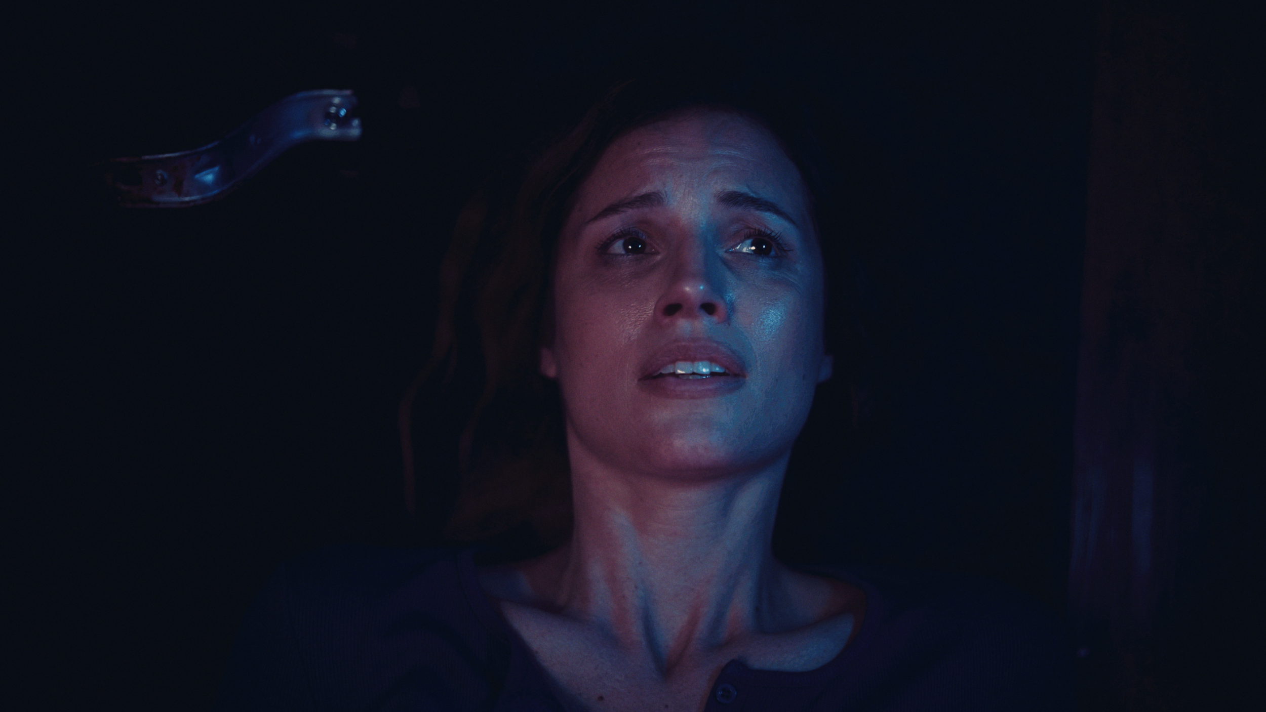 A woman lying down with a frightened or distressed expression, dark background, blue lighting, with a metallic object or piece of machinery near her head.
