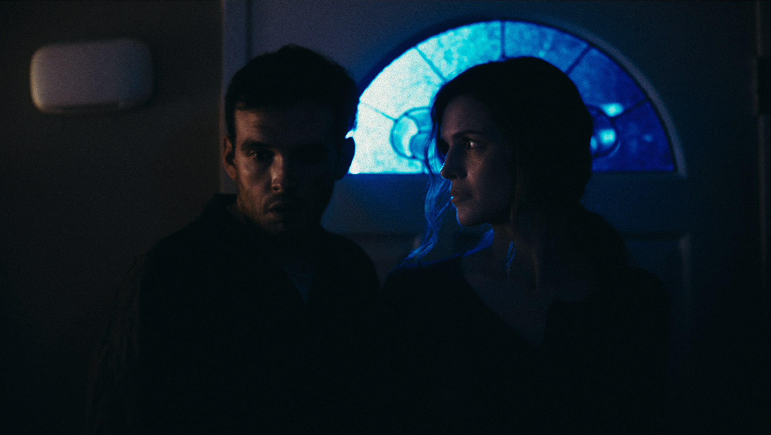 A man and woman sitting in a dimly lit room with a blue-lit window behind them, looking in different directions.