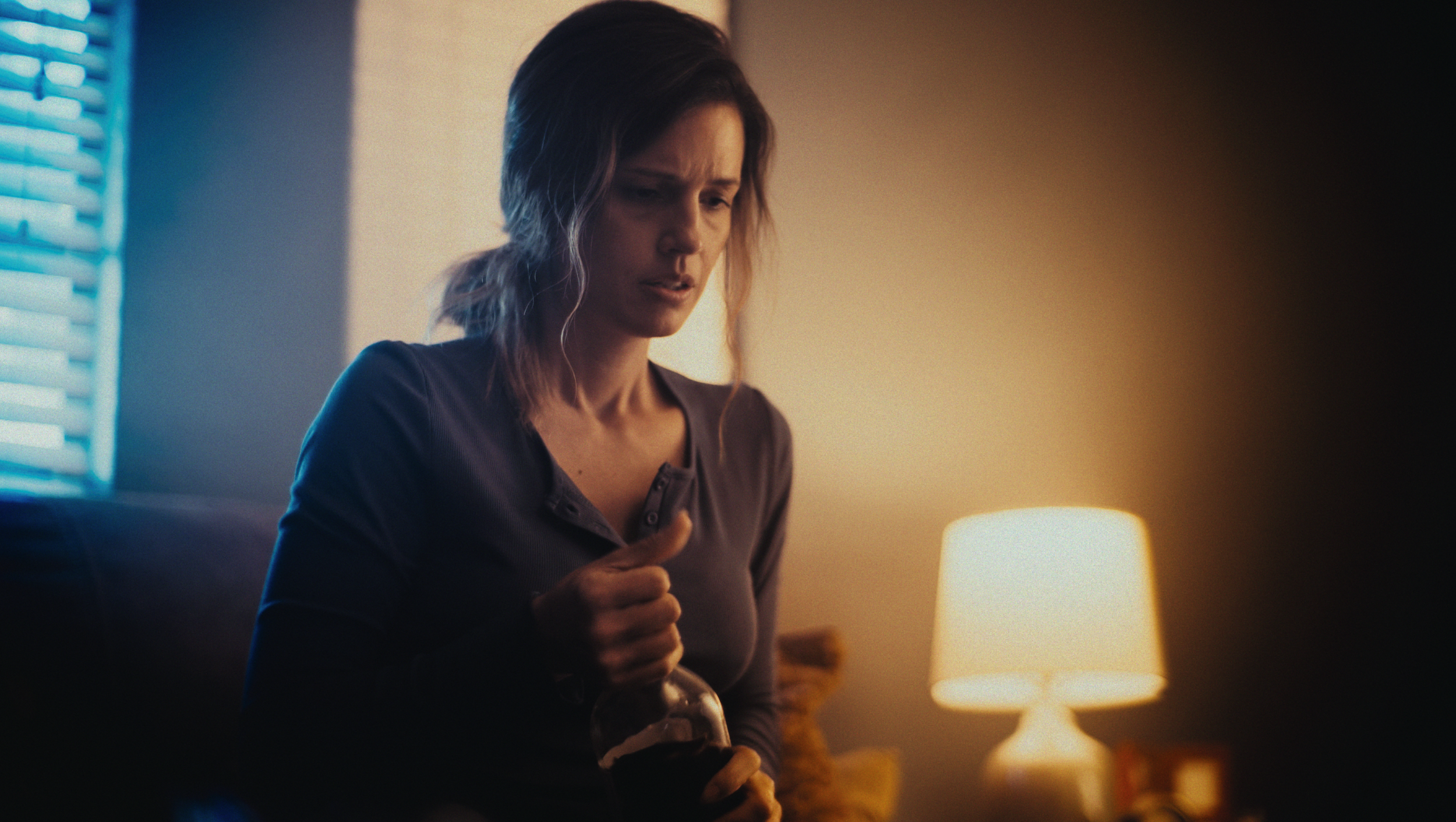 A woman with disheveled hair, wearing a dark shirt, sits in a dimly lit room holding a glass bottle, with a concerned or distressed expression.