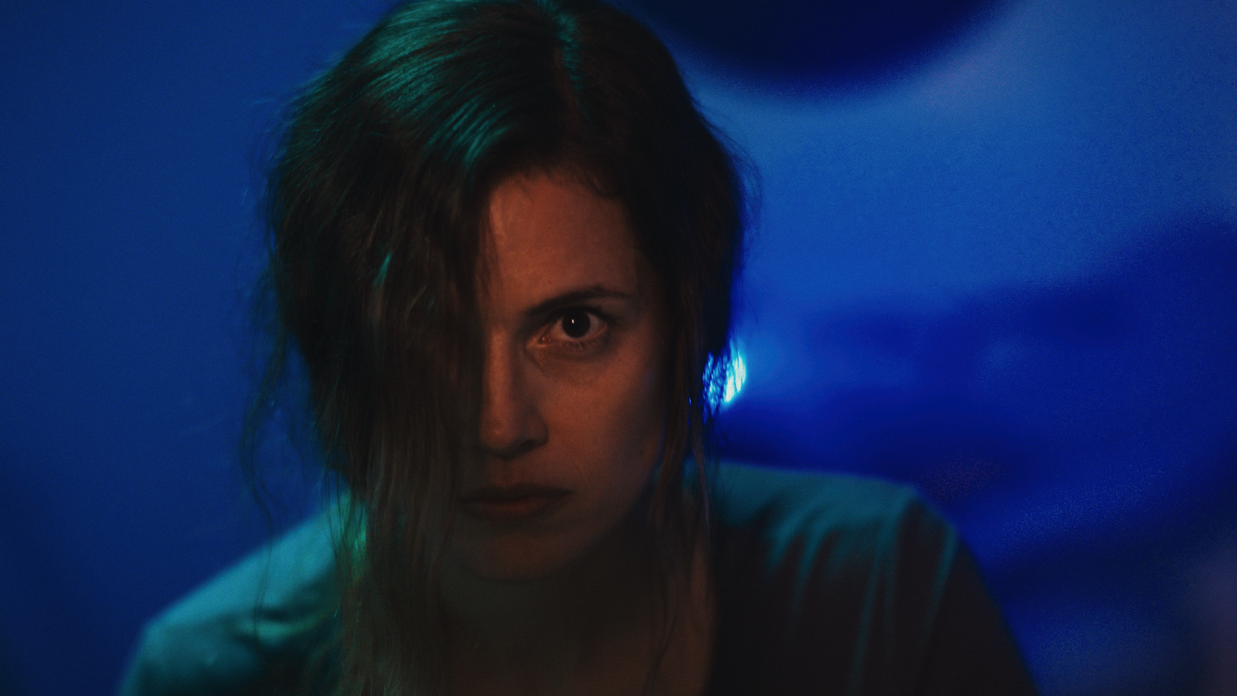 A woman with wet hair partially covering her face, staring intently at the camera, with blue and green lighting creating a dramatic atmosphere.