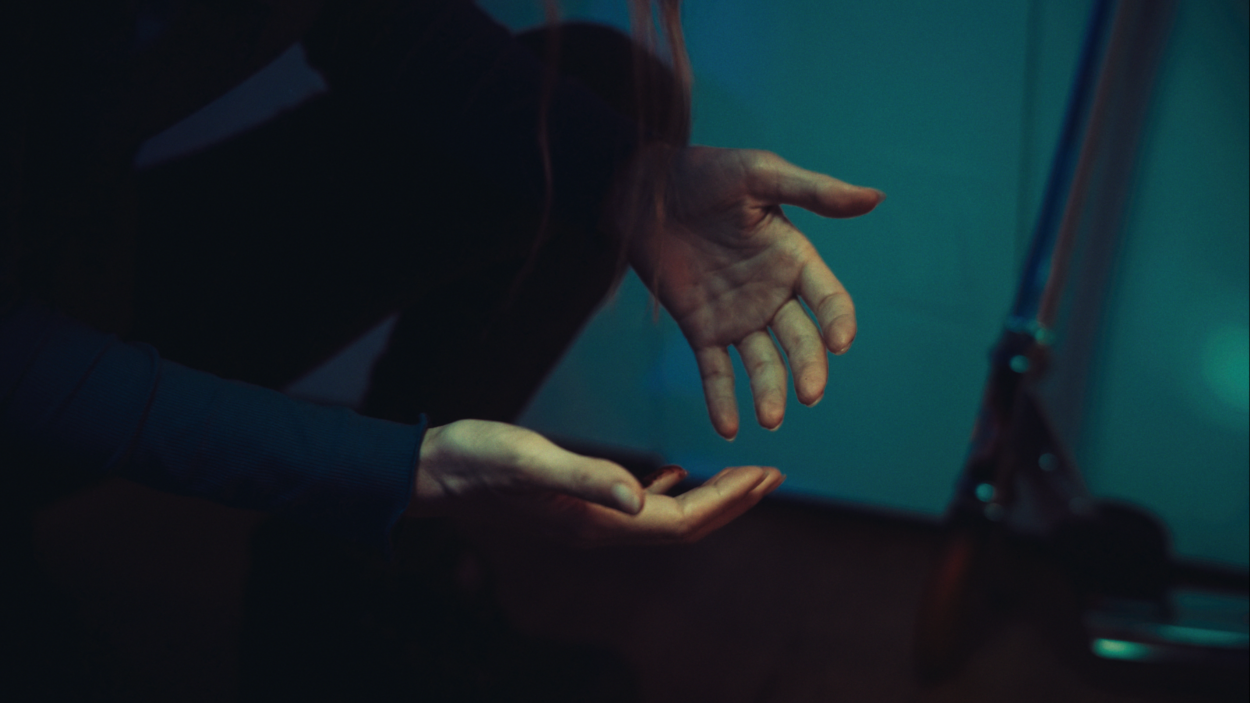 Close-up of two hands, one reaching out and the other open, under low lighting with a blue-green background.