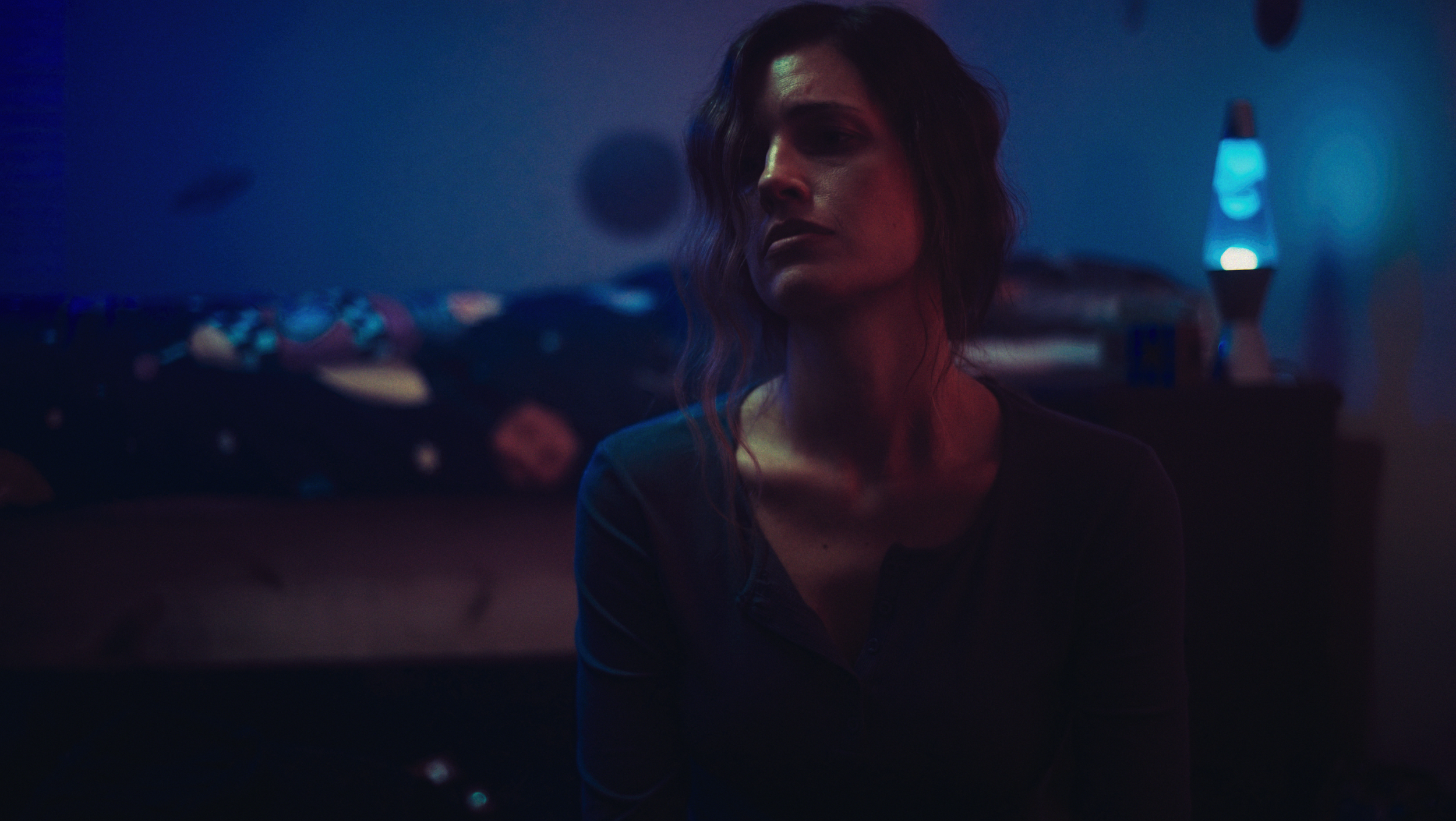 A young woman with wavy hair sits in a dimly lit room illuminated by blue and purple neon lights, with a bed and a lava lamp in the background.