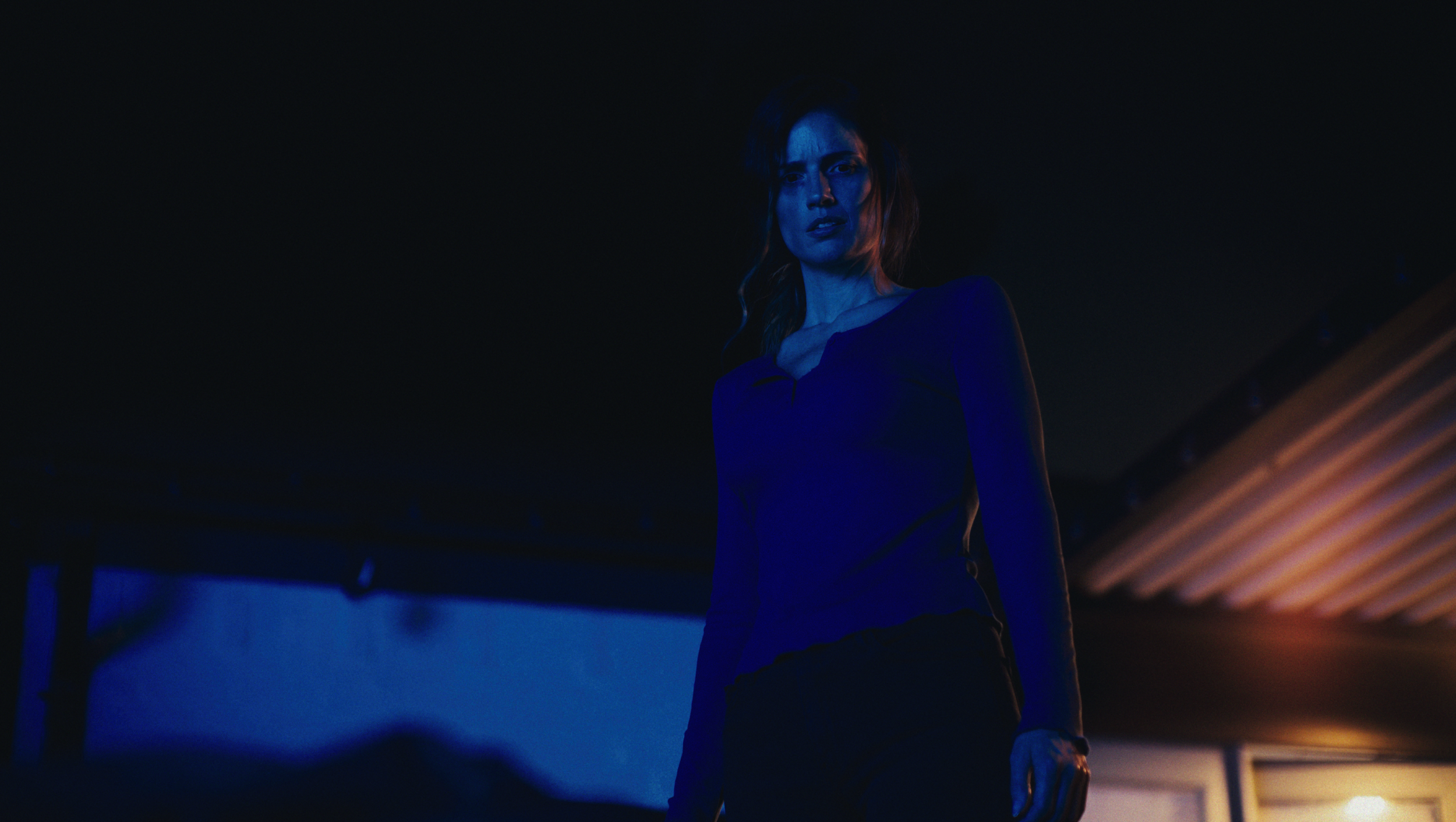 A woman with long hair, illuminated by blue and orange lighting, stands in a dark room with a serious or intense expression.