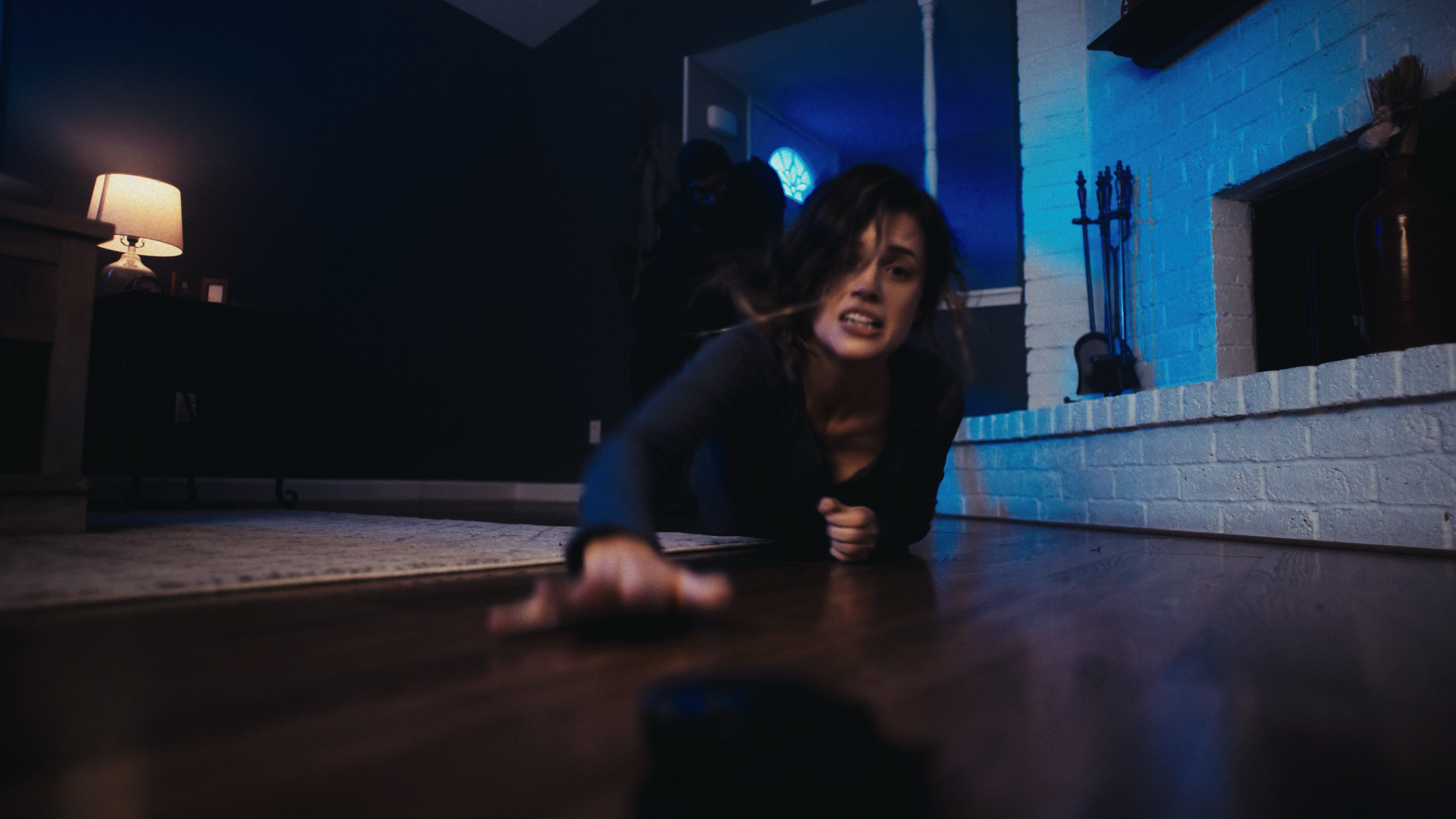 Woman on the floor with a distressed expression, reaching out toward the camera, with a man standing behind her in a dimly lit living room near a fireplace.