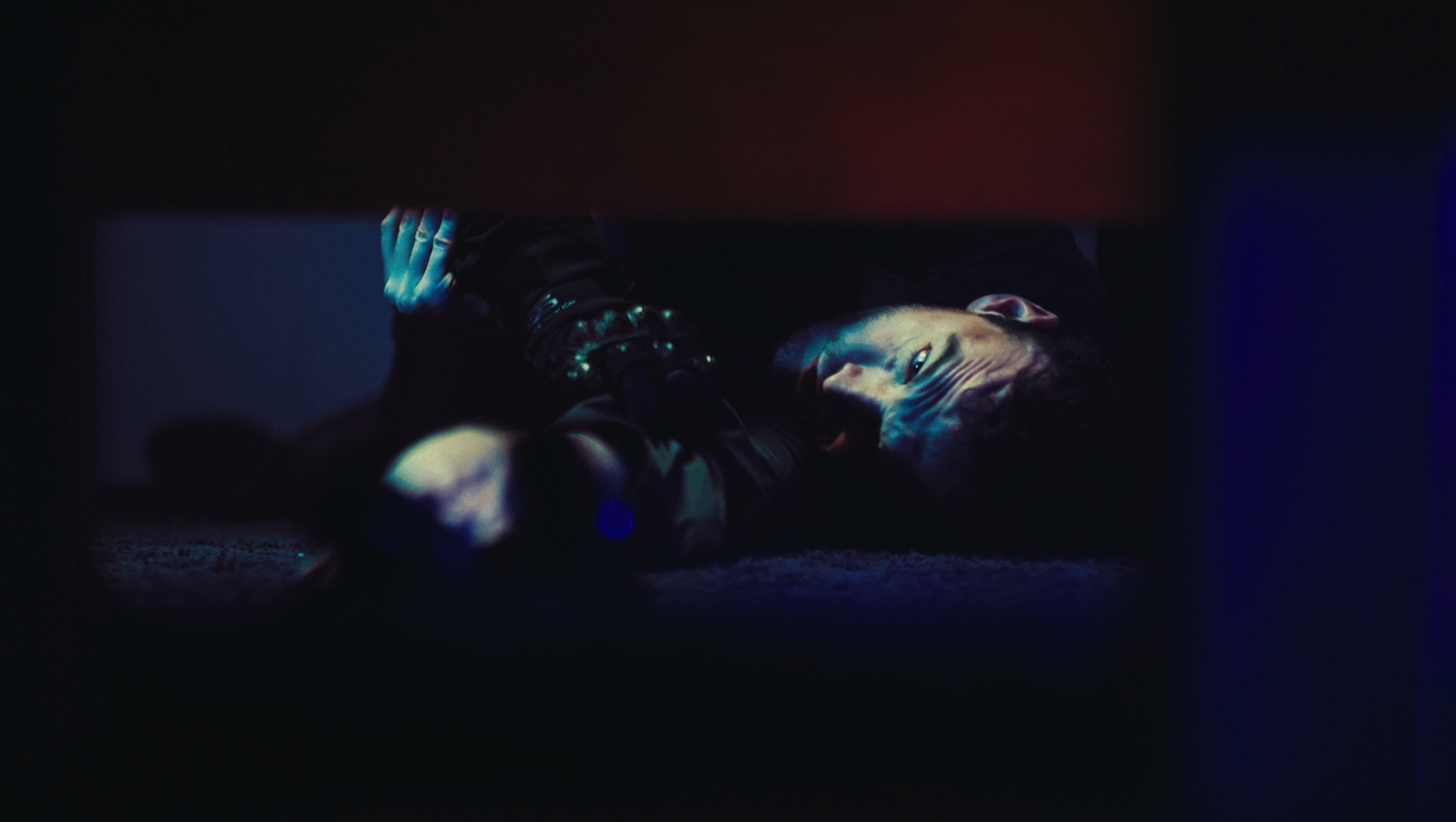 A man lying on the floor under a piece of furniture, illuminated by blue and purple lights, with a distressed or intense expression.