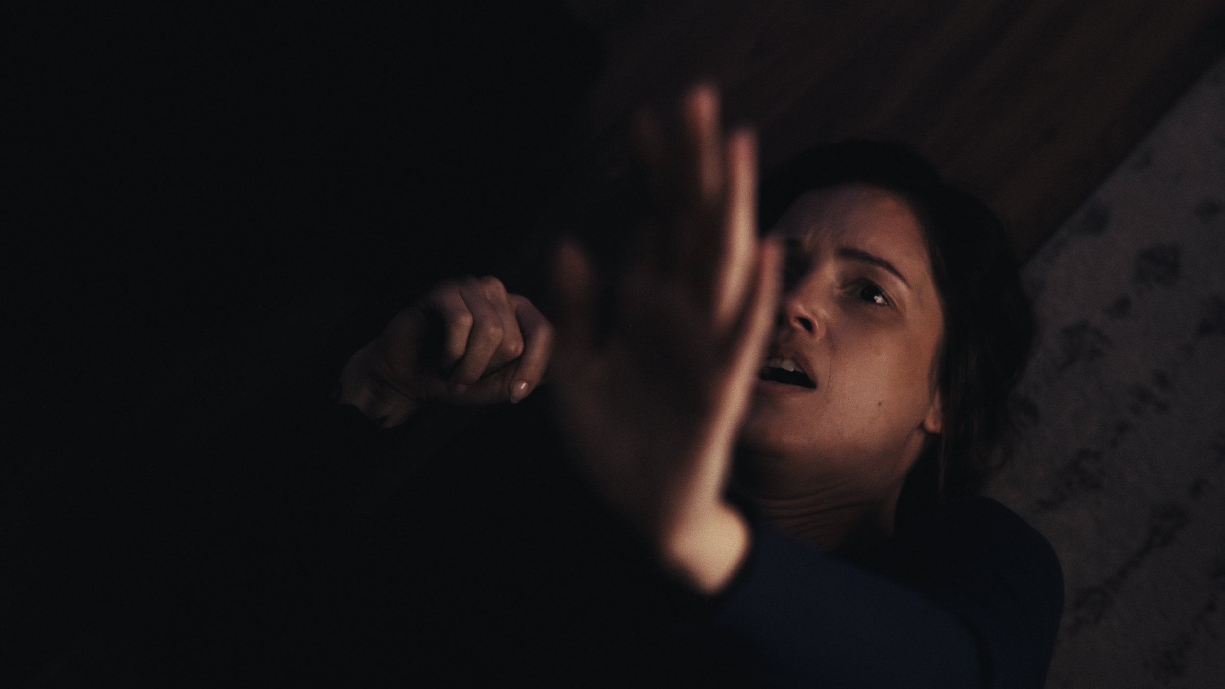 A woman lying on the floor with a distressed expression, reaching out with her hand towards a dark figure.