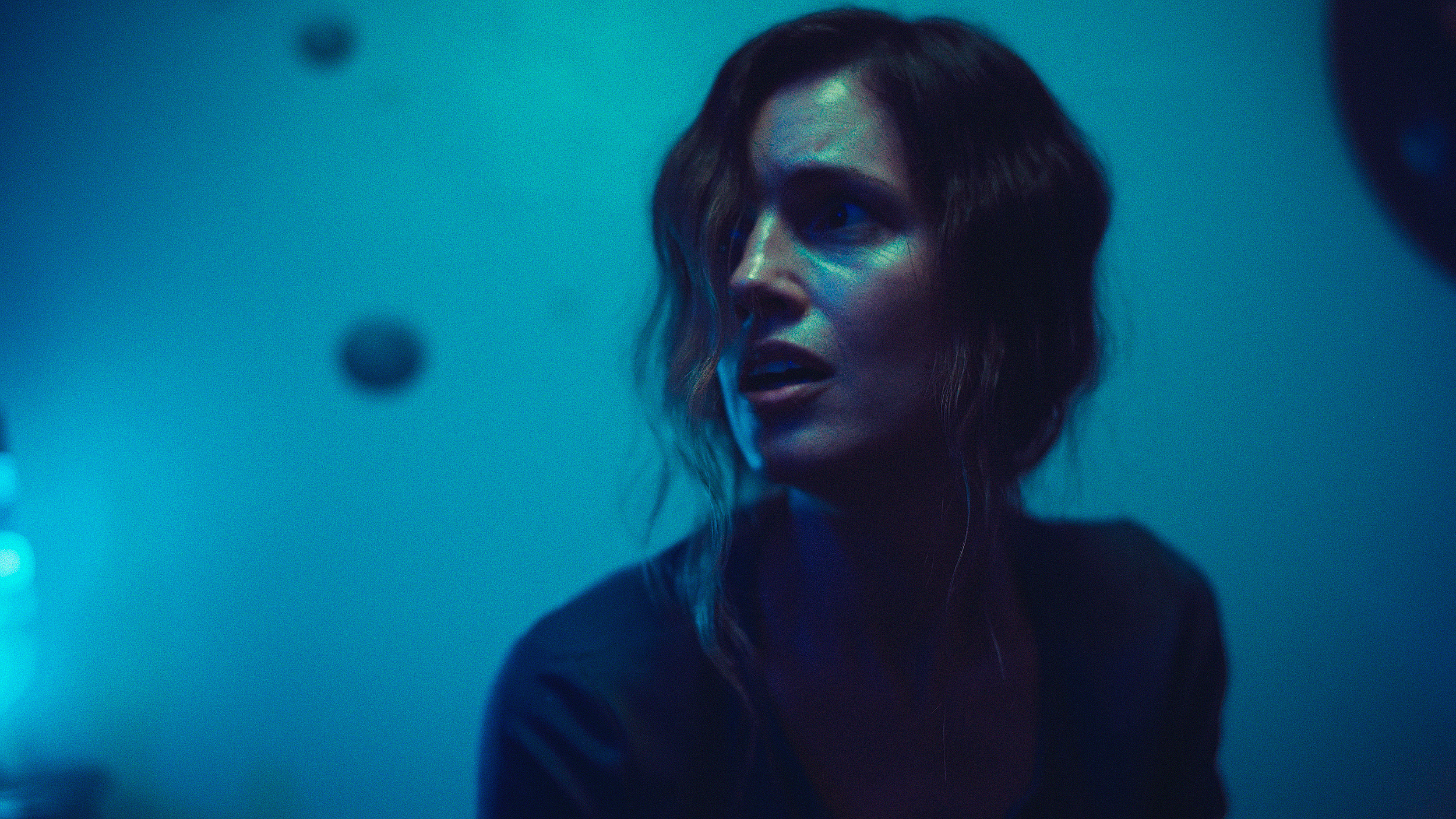 A woman with short, wavy hair and makeup looking to the side, in a dimly lit room with blue lighting.