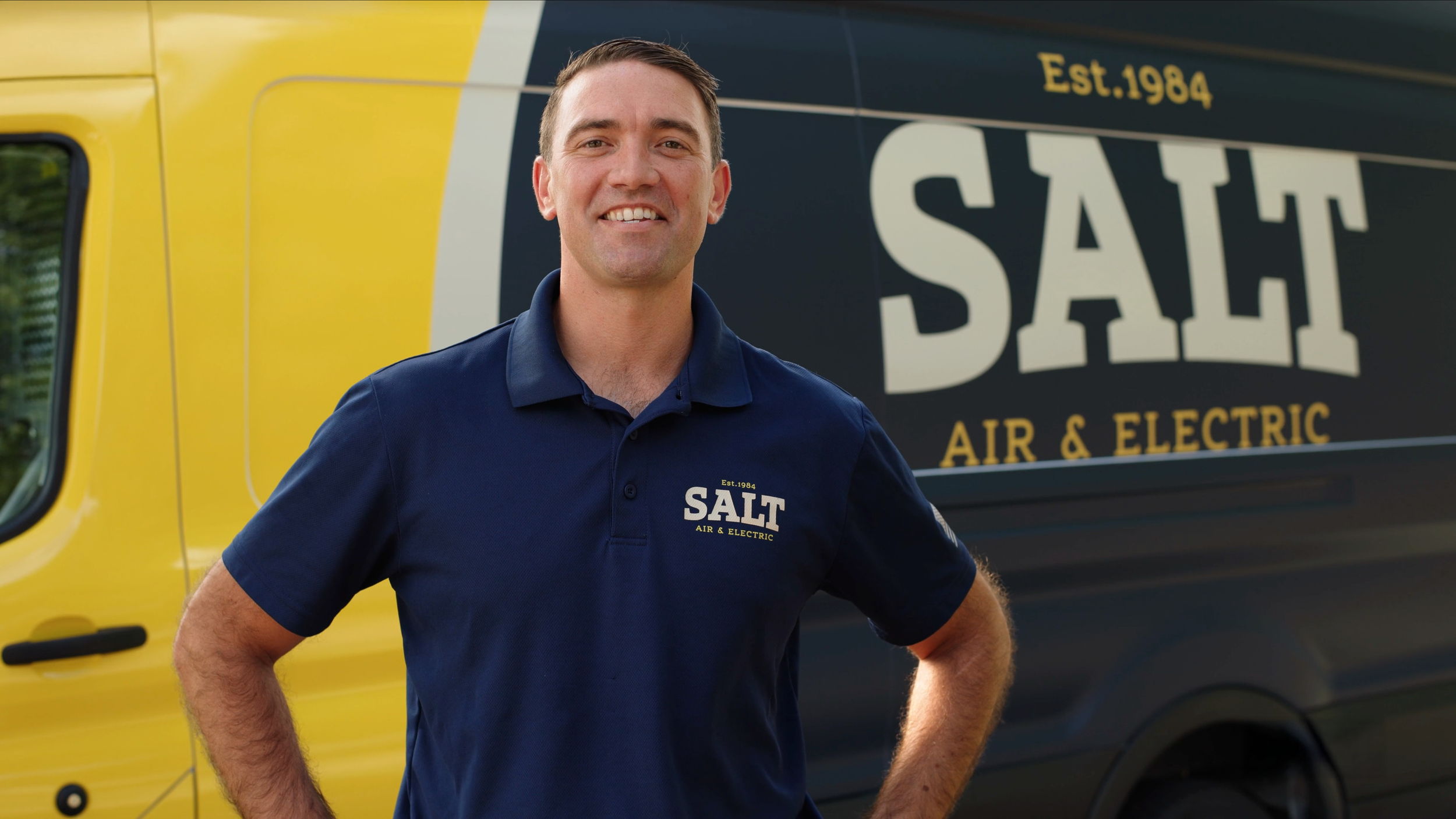 A man wearing a navy blue polo shirt with the SALT Air & Electric logo standing in front of a yellow and black SALT Air & Electric service van.