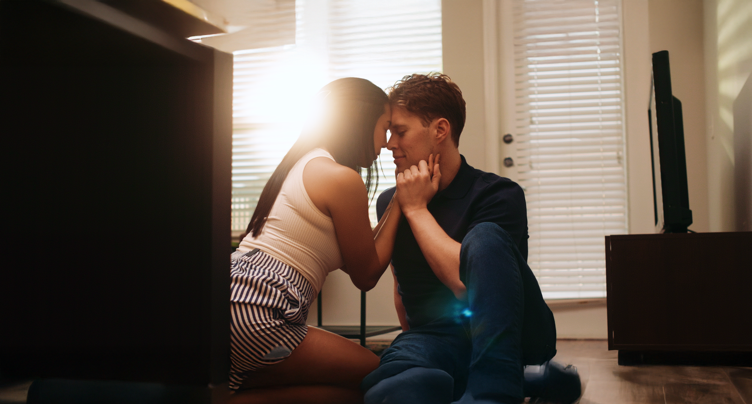 A young couple sitting on the floor indoors, with their foreheads touching and eyes closed, embracing each other lovingly, backlit by sunlight coming through the blinds.