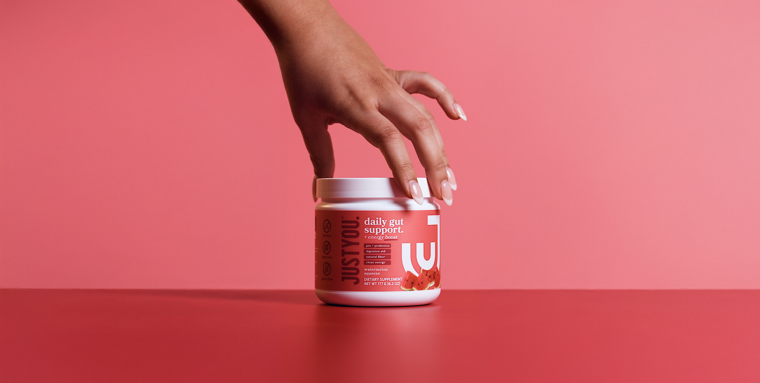 A person's hand resting on a pink surface near a white container of juicePlus dietary supplement with a pink background.