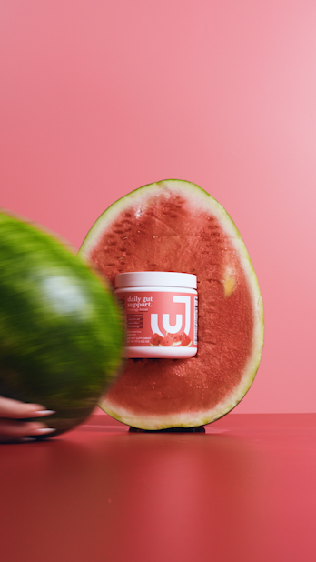 A container of beauty or skincare product placed inside a cut watermelon half against a pink background, with a blurry watermelon in the foreground.