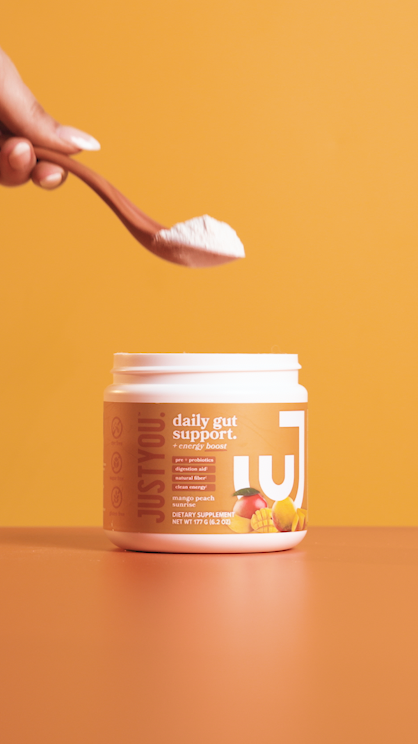 A hand holding a wooden spoon with white powder above a container labeled "Justed™ daily gut support" against an orange background.
