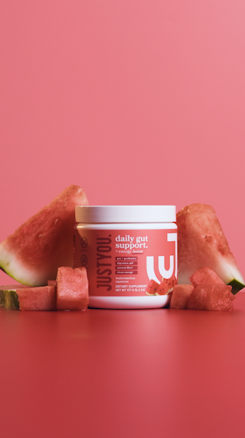 A container of Just You! daily gut support supplement surrounded by slices of watermelon and watermelon cubes on a pink background.