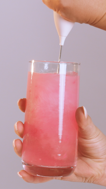 Hand holding a glass of pink beverage being decorated with a white piping tool.