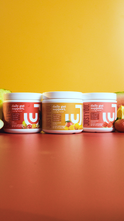 Three containers of Just You daily gut support supplements, with a yellow, pink, and red label, placed on a pink surface against a yellow background, with parts of fruits visible on each side.